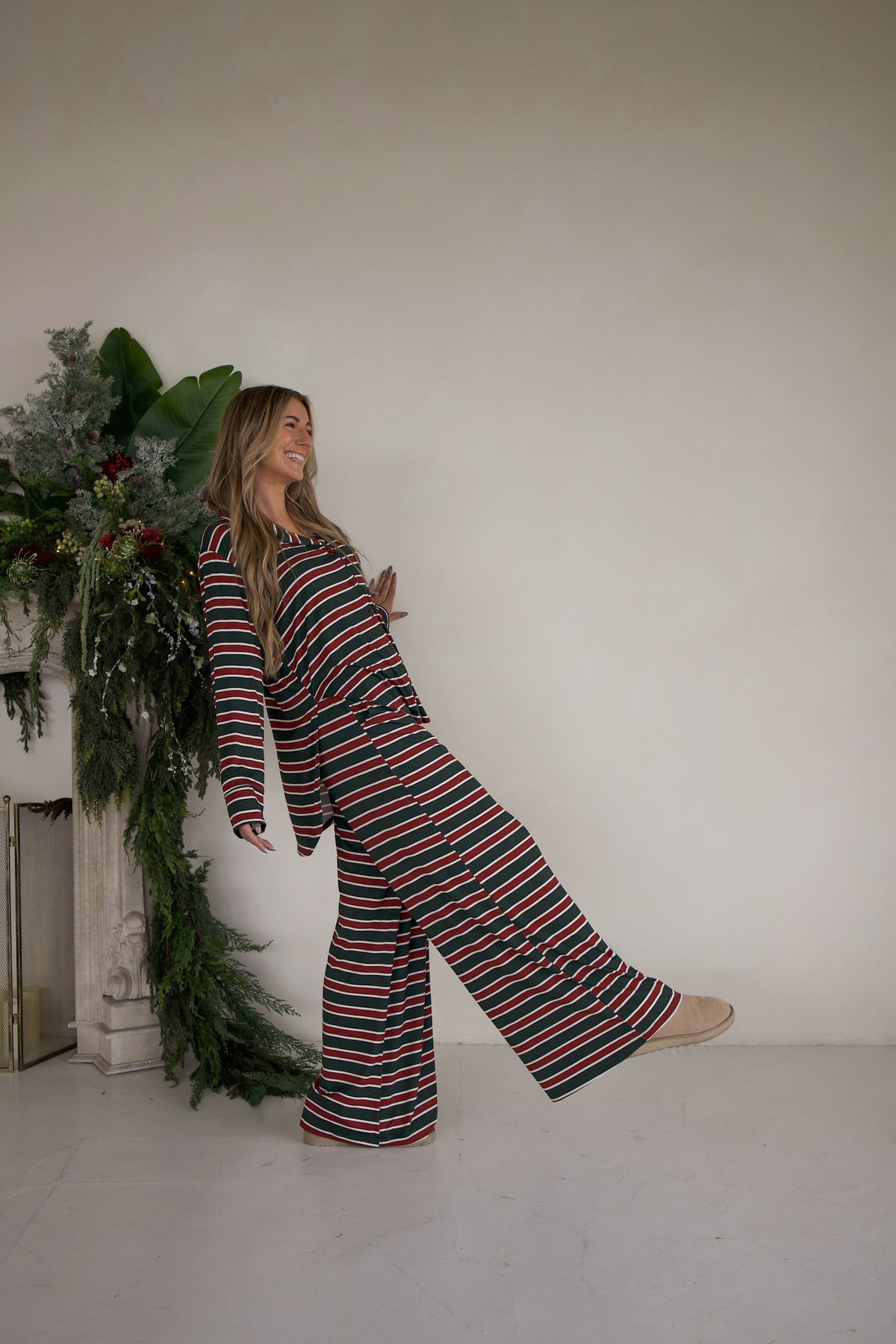 A smiling woman wears a red, green, and white Women’s Bamboo Button Up Lounge Set | Holiday Stripe with wide-leg pants while leaning against a fireplace decorated with greenery.