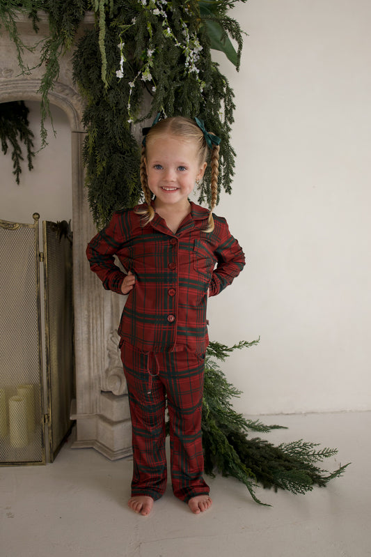 A young girl stands with hands on her hips wearing a Girl’s Bamboo Button Up Lounge Set | Christmas Plaid, a long-sleeved, button-up pajama set.