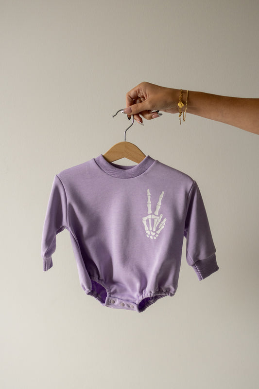 A hand holds a hanger with the forever french baby Sweatshirt Romper | Enchanted Skelly, crafted from 100% cotton in a lavender midnight shade and featuring a white skeleton hand making a peace sign printed on the chest.
