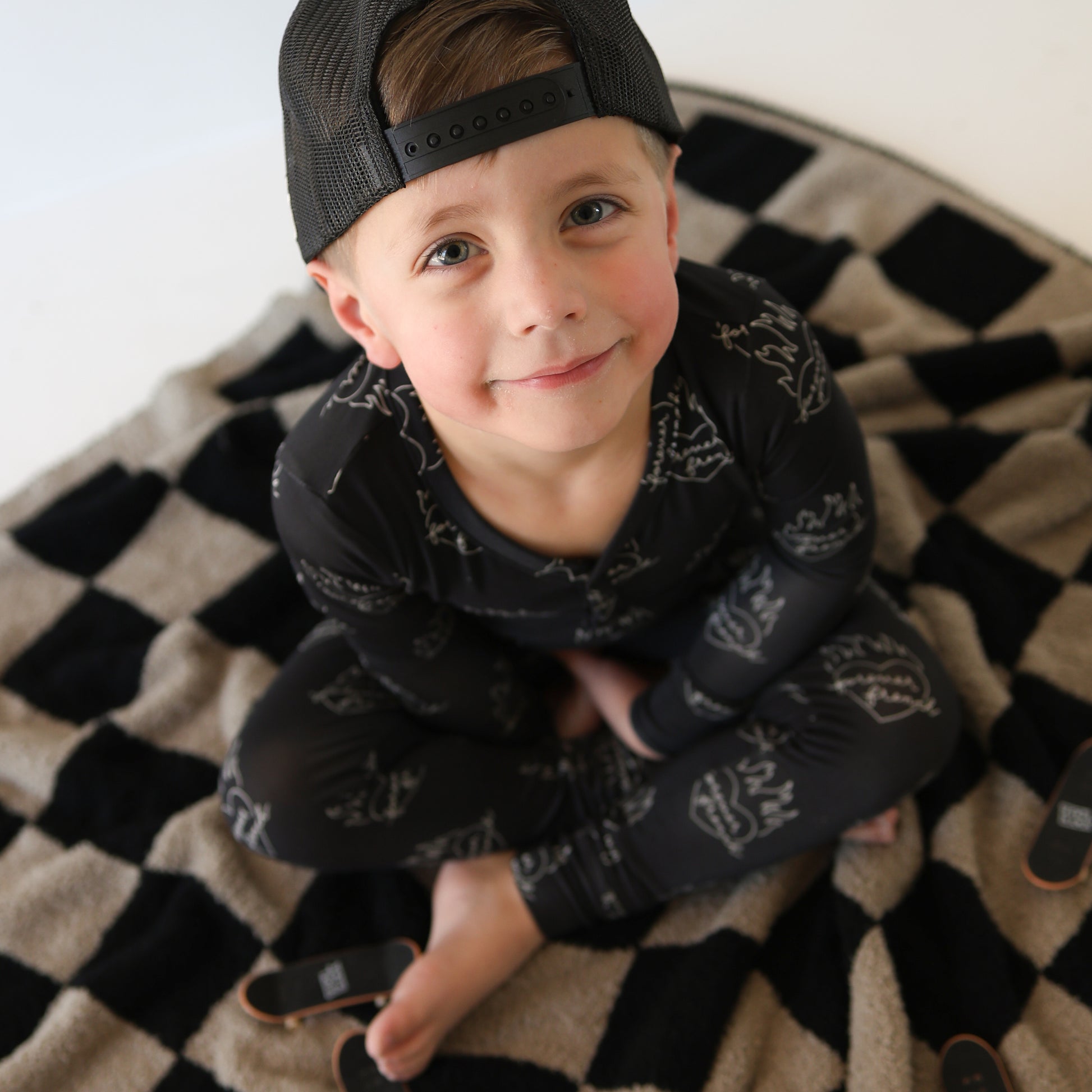 A young boy in forever french baby's Bamboo Two Piece Pajamas | Flaming Hearts sits cross-legged on a black and beige checkered blanket, smiling up at the camera with a black cap worn backwards. Two miniature skateboards are beside him.