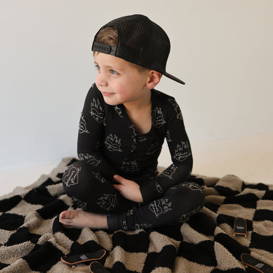 A young child in Forever French Baby’s Bamboo Two Piece Pajamas | Flaming Hearts sits cross-legged on a black and beige patterned blanket, smiling and looking to the side.