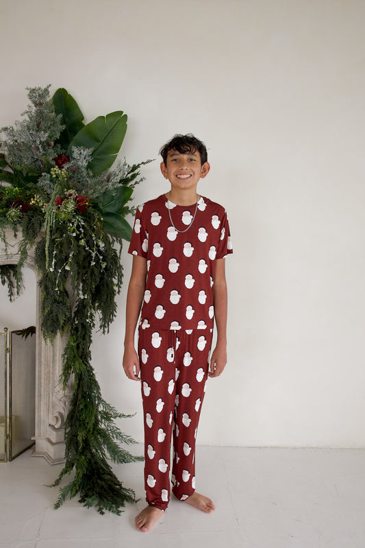 A smiling boy wears Youth Straight Leg Bamboo Pajamas | Vintage Santa, a matching set with a short-sleeved top and pants featuring a dark red background and a white Santa Claus pattern.