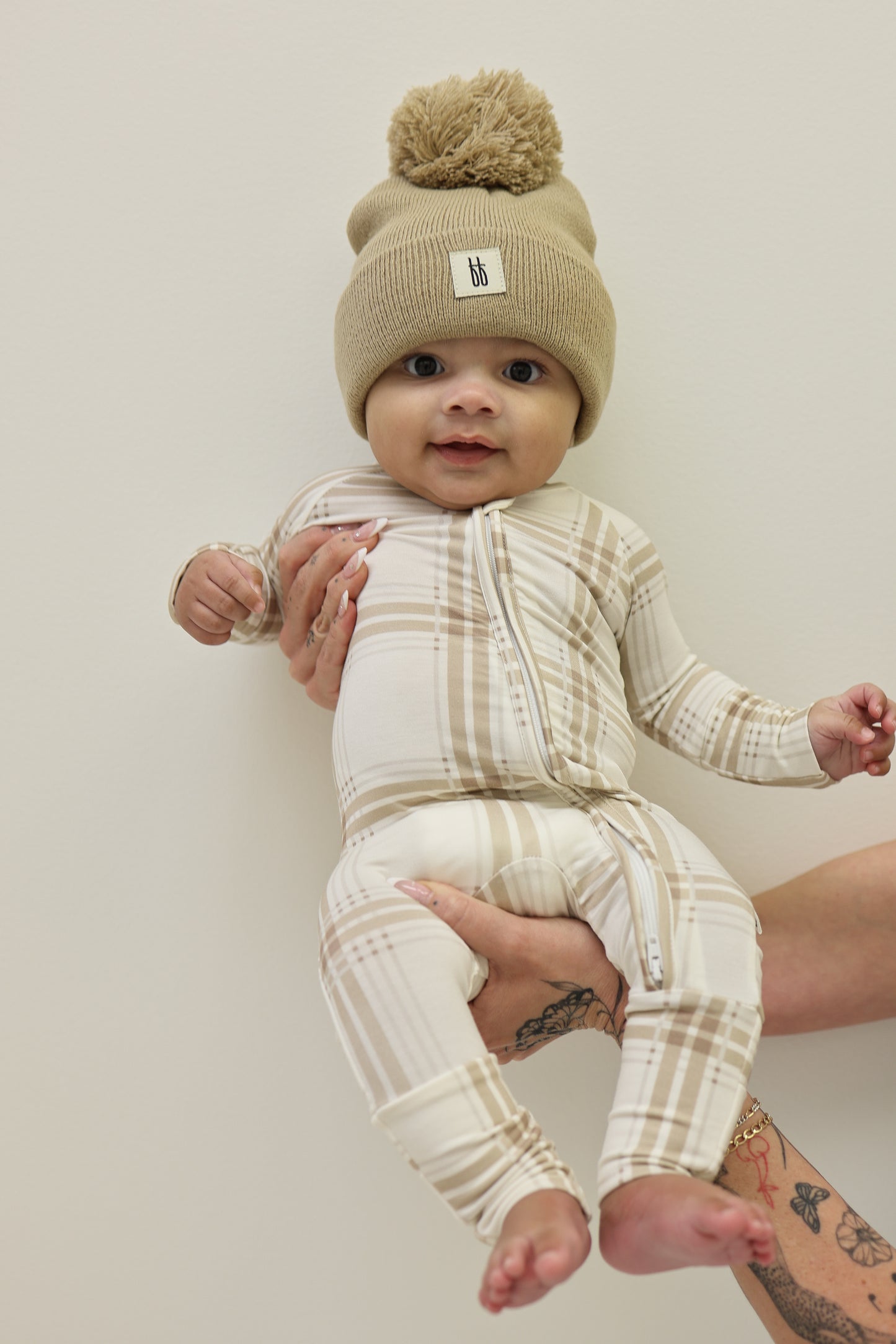A baby in forever french baby's Bamboo Zip Pajamas | Ellis Plaid—a beige plaid onesie—plus a tan knit hat with pom-pom, is held by a tattooed adult against a plain, light background.
