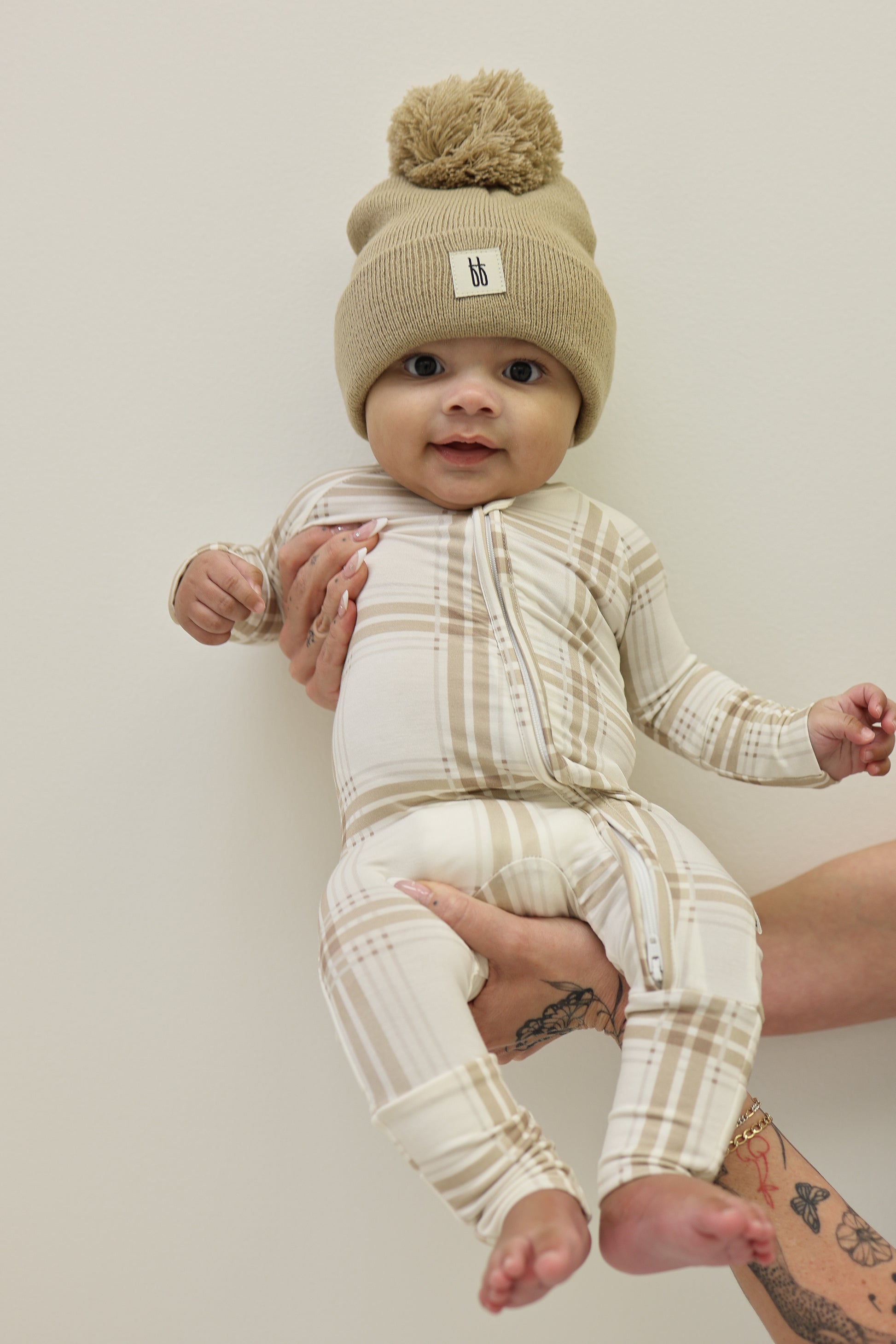 A baby in forever french baby's Bamboo Zip Pajamas | Ellis Plaid—a beige plaid onesie—plus a tan knit hat with pom-pom, is held by a tattooed adult against a plain, light background.