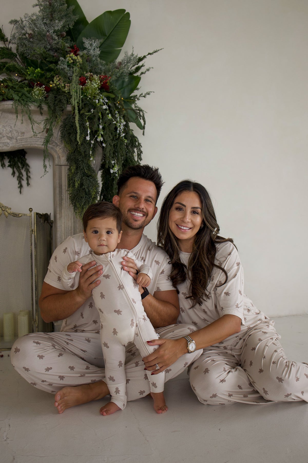 A family of three sitting together in matching Bamboo Zip Pajamas | Gingersnap, featuring a breathable brown gingerbread man print.