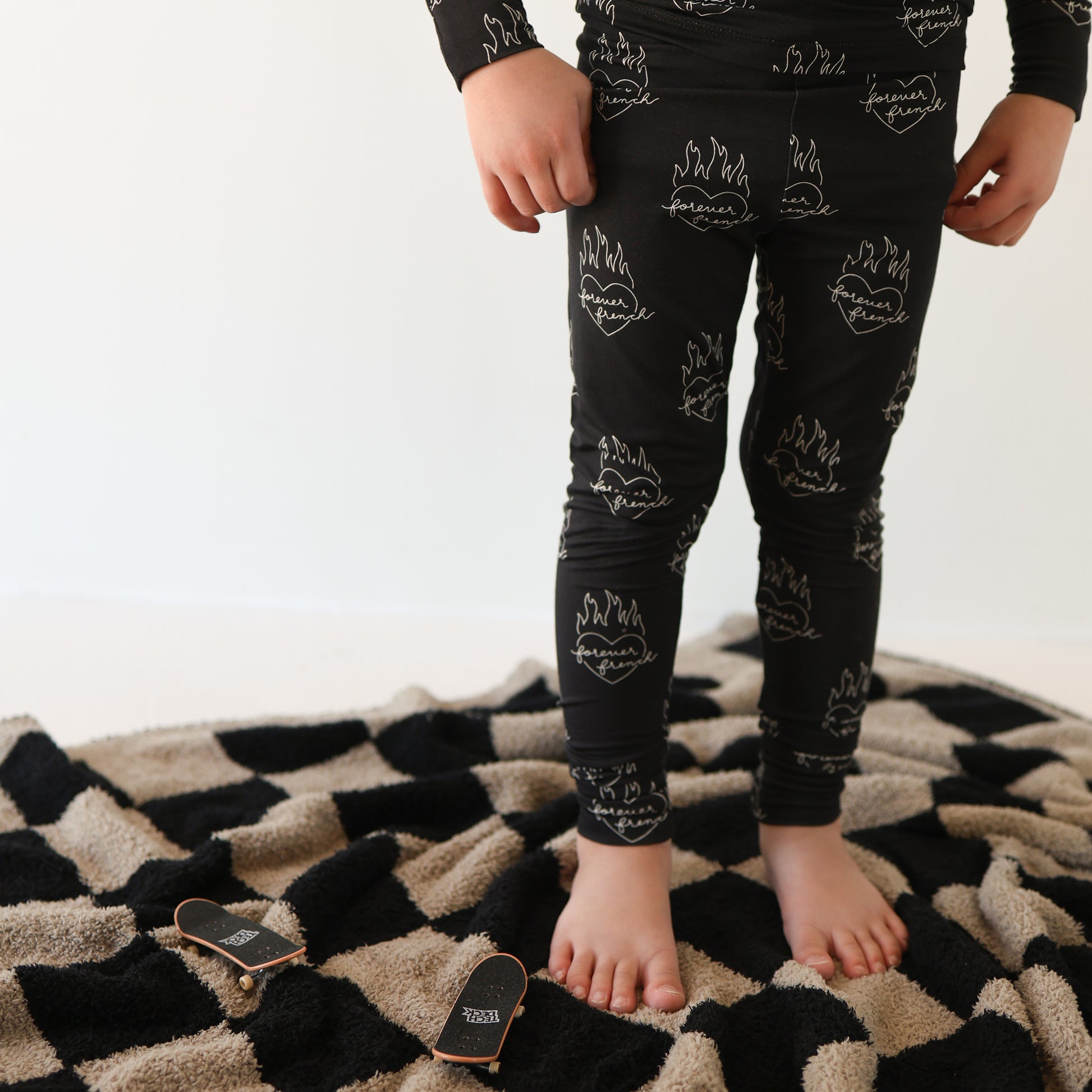A child stands barefoot on a black and beige checkered rug, wearing forever french baby Bamboo Two Piece Pajamas | Flaming Hearts. Two small wooden finger skateboards with black tops and labels lie nearby.