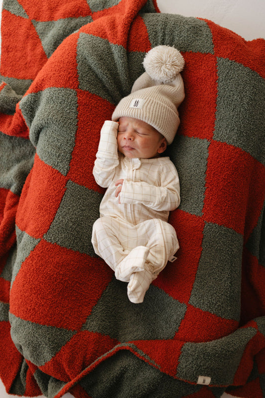 A newborn baby sleeps peacefully on the forever french baby's "Retro Christmas | Plush Blanket," its red and green checkered pattern evoking festive nostalgia. The baby is cozily dressed in a light-colored striped sleeper and a beige knit hat adorned with a fluffy pom-pom.