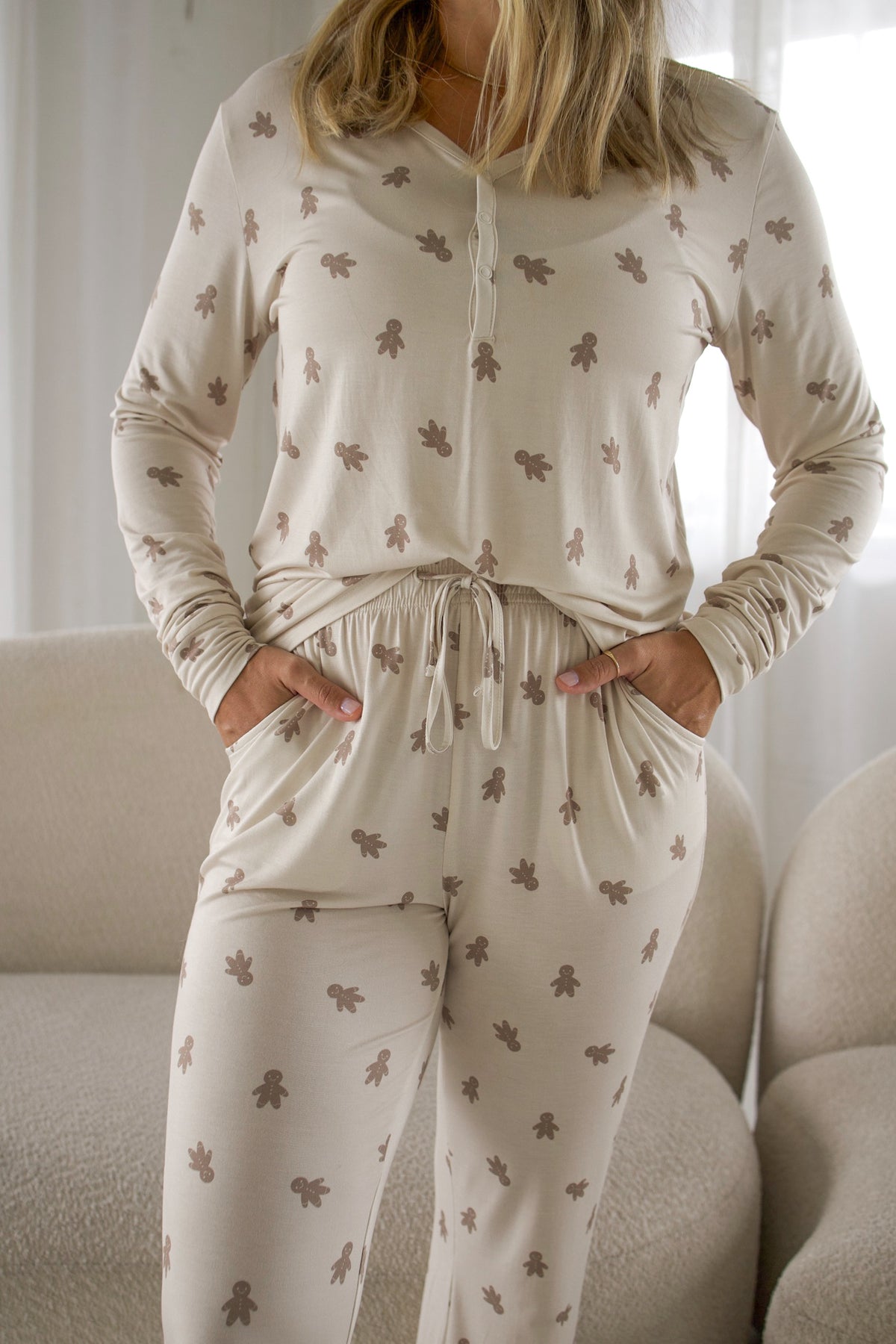 A woman wearing tan long-sleeve Women’s Long Sleeve Bamboo Pajama | Gingersnap with a matching gingerbread man print, her hands tucked into the pockets of the pants.
