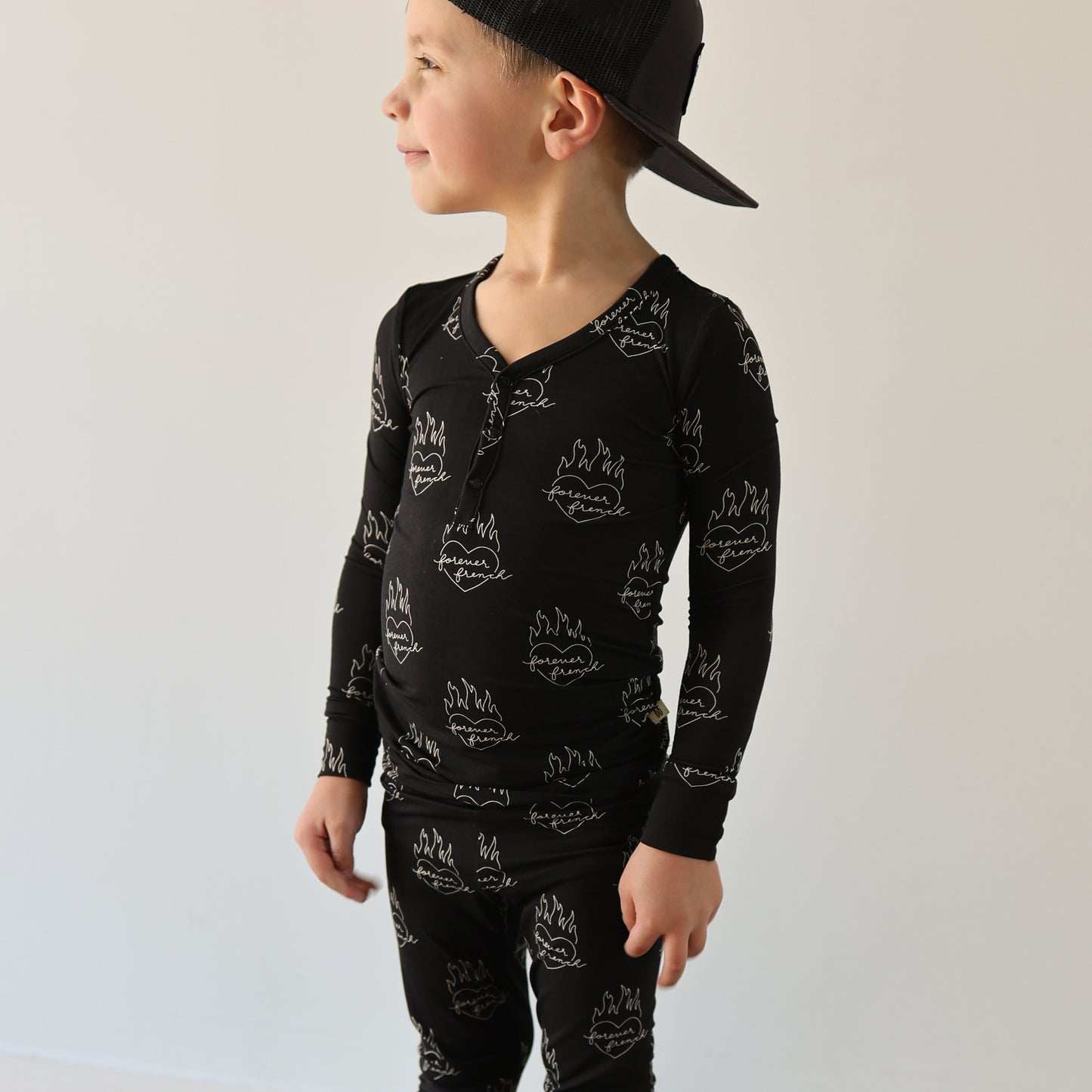 A young child in forever french baby’s Bamboo Two Piece Pajamas | Flaming Hearts—a black, hypoallergenic set with white flame and smiley faces—stands against a light wall, looking sideways and wearing a backward black cap.