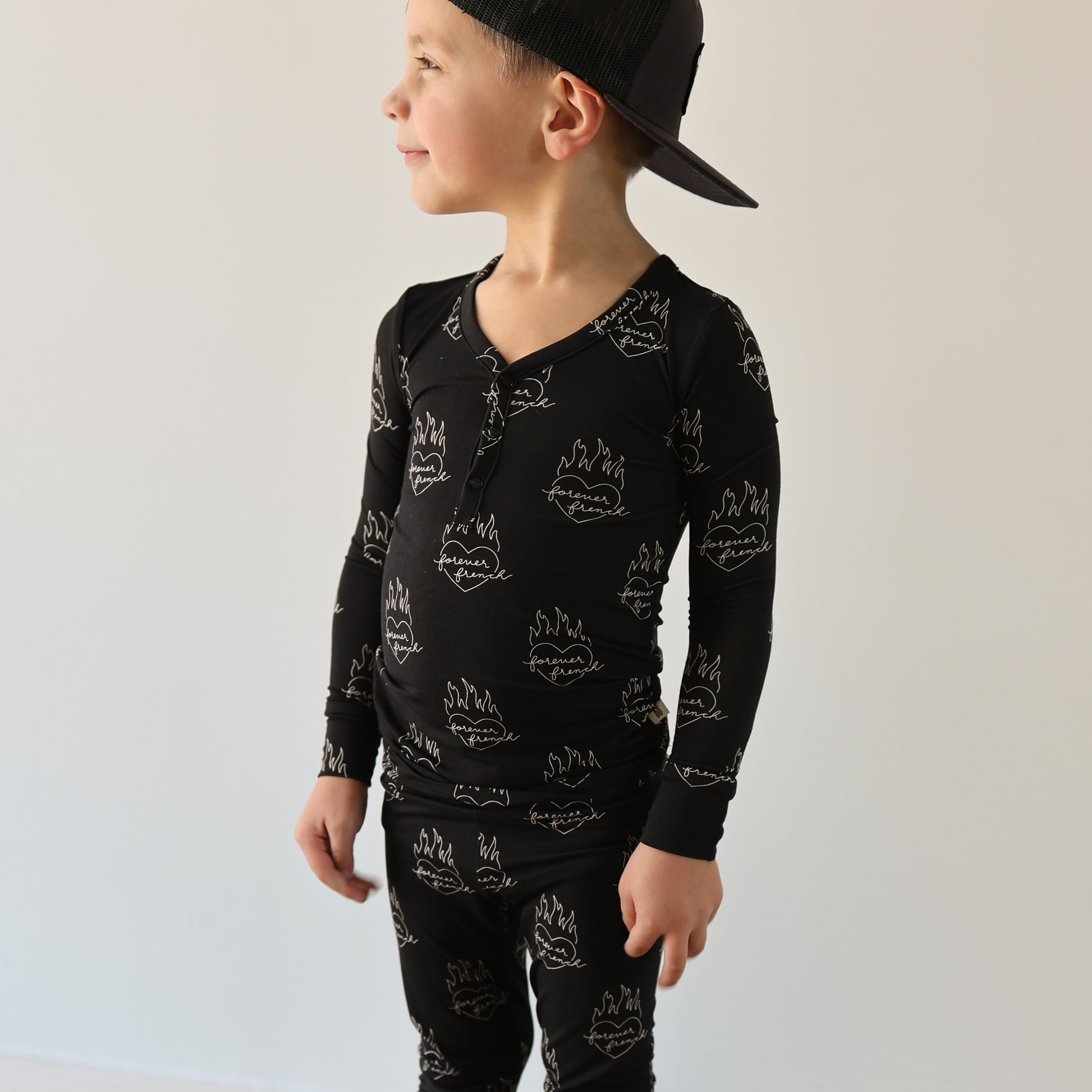 A young child in forever french baby’s Bamboo Two Piece Pajamas | Flaming Hearts—a black, hypoallergenic set with white flame and smiley faces—stands against a light wall, looking sideways and wearing a backward black cap.