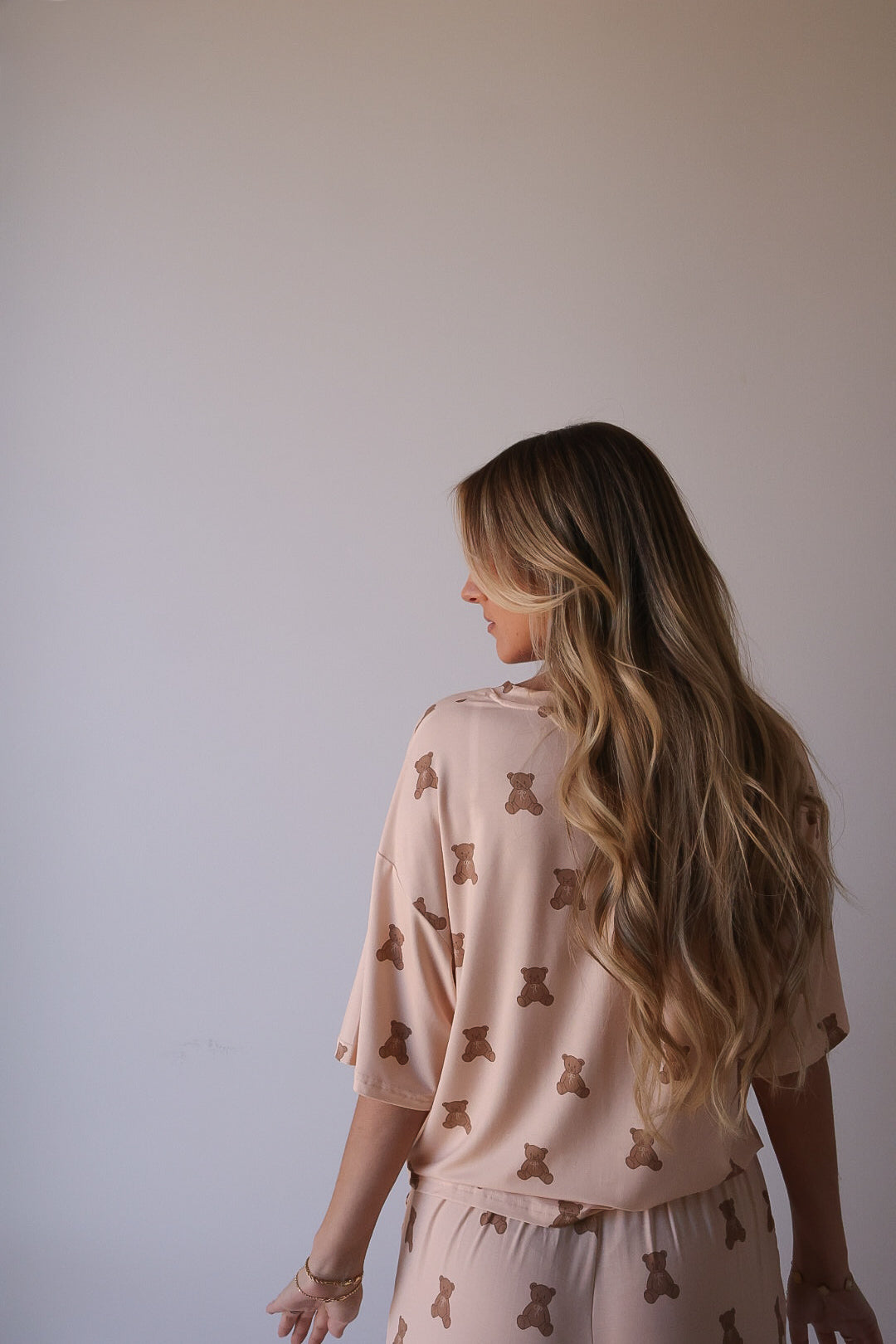 A woman with long, wavy blonde hair faces away, wearing the forever french baby Women's Boxy Tee Lounge Set | Lottie—a cozy, hypo-allergenic bamboo set featuring a teddy bear print on light beige against a plain wall.