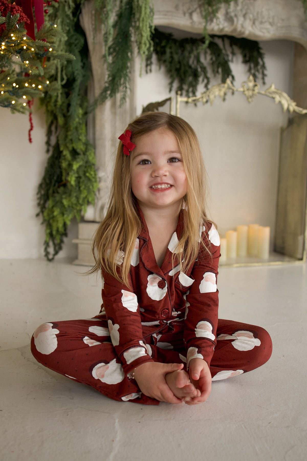 A young girl sits cross-legged wearing a Girl’s Bamboo Button Up Lounge Set | Vintage Santa patterned with Santa Claus faces.