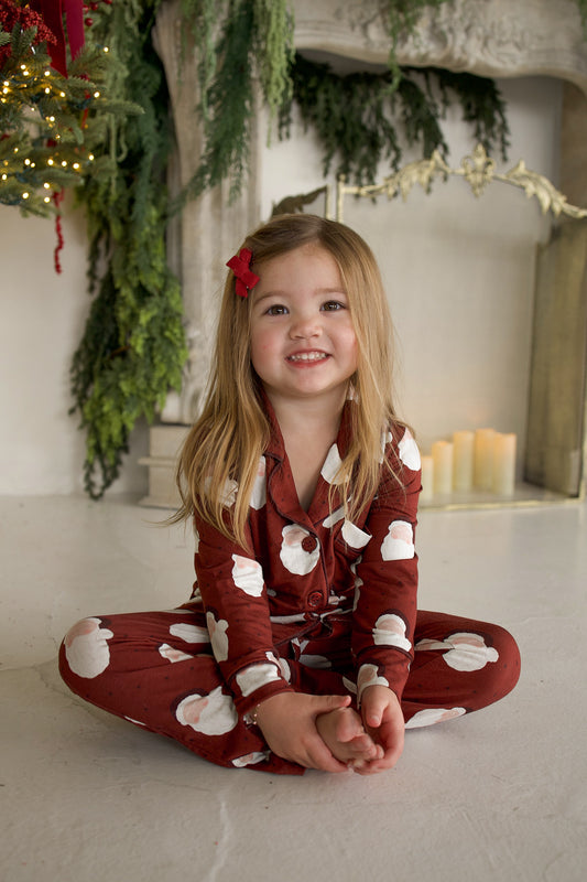 A young girl sits cross-legged wearing a Girl’s Bamboo Button Up Lounge Set | Vintage Santa patterned with Santa Claus faces.