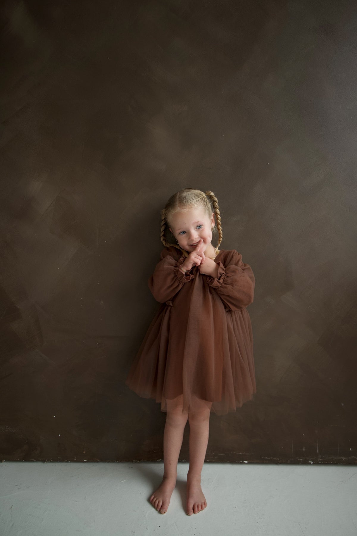 A young girl with braided hair wears a brown Long Sleeve Tutu Dress | Cocoa and holds her fingers to her lips.