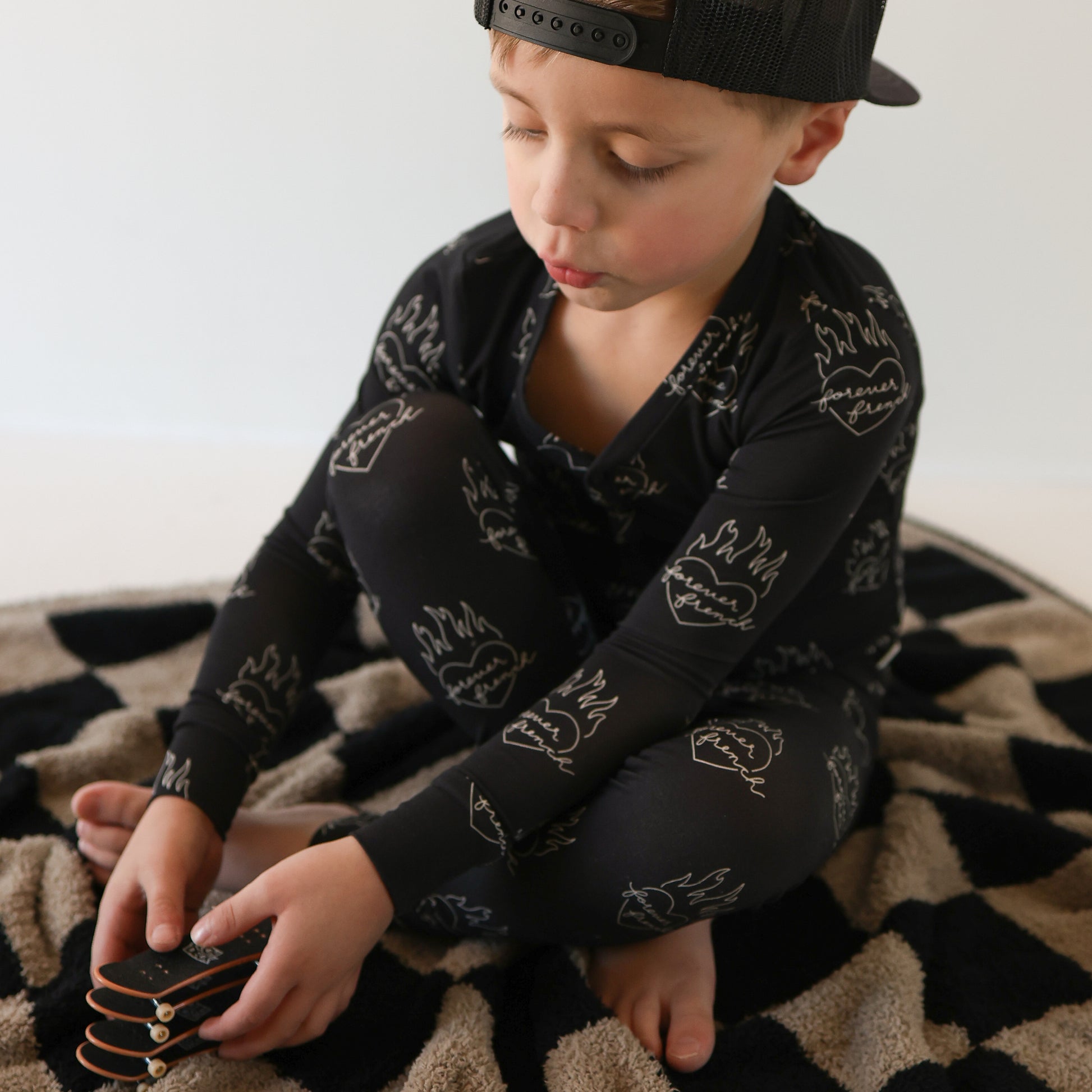 A young boy in forever french baby’s Bamboo Two Piece Pajamas | Flaming Hearts—black, flame-print, hypoallergenic sleepwear—sits cross-legged on a patterned rug, focused as he stacks small black blocks.