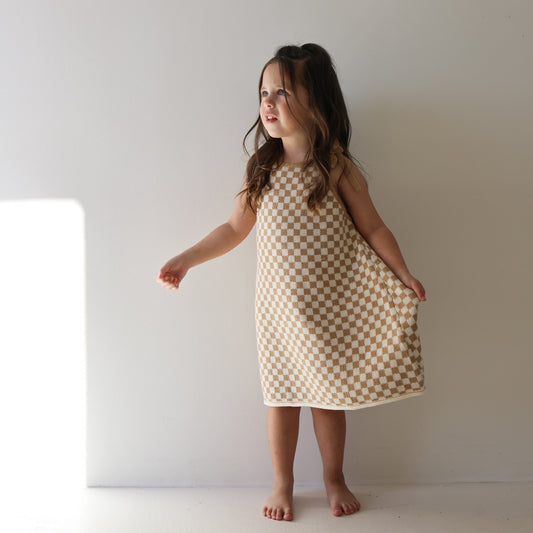 A young girl with long brown hair stands barefoot against a white wall, wearing the Knit Dress | Latte Check from forever french baby, gazing softly to her right as sunlight filters in from the left.