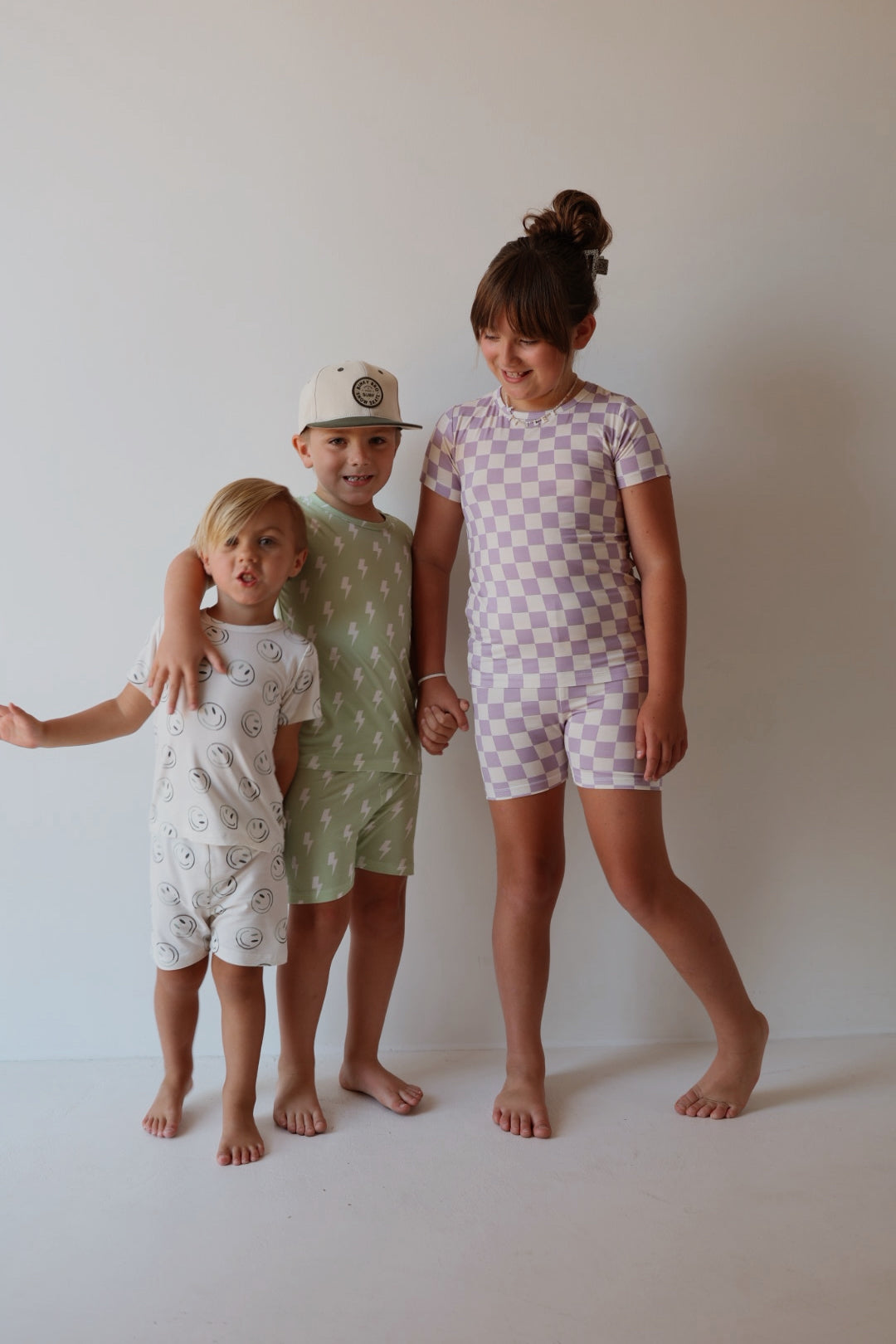 Three barefoot children stand against a light background. The youngest wears a white romper with smiley faces, the middle child wears bamboo pajamas and a white hat, and the oldest wears the Bamboo Short Set | Grape Soda by forever french baby. They are smiling and holding hands.