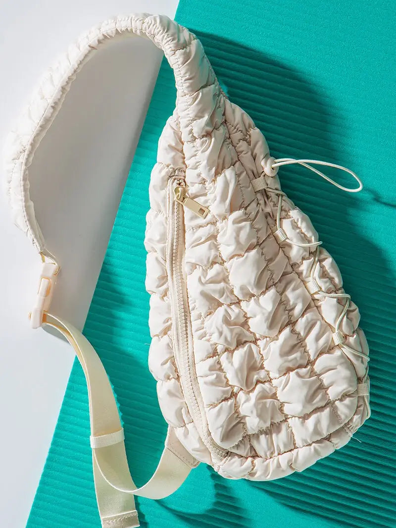 The urban daizy Quilted Sling Bag, showcased against a teal and white background, is a white, ruched design accessory that boasts a zipper running down the center and an adjustable strap. Perfect for everyday use, this stylish bag combines functionality with elegant fashion.