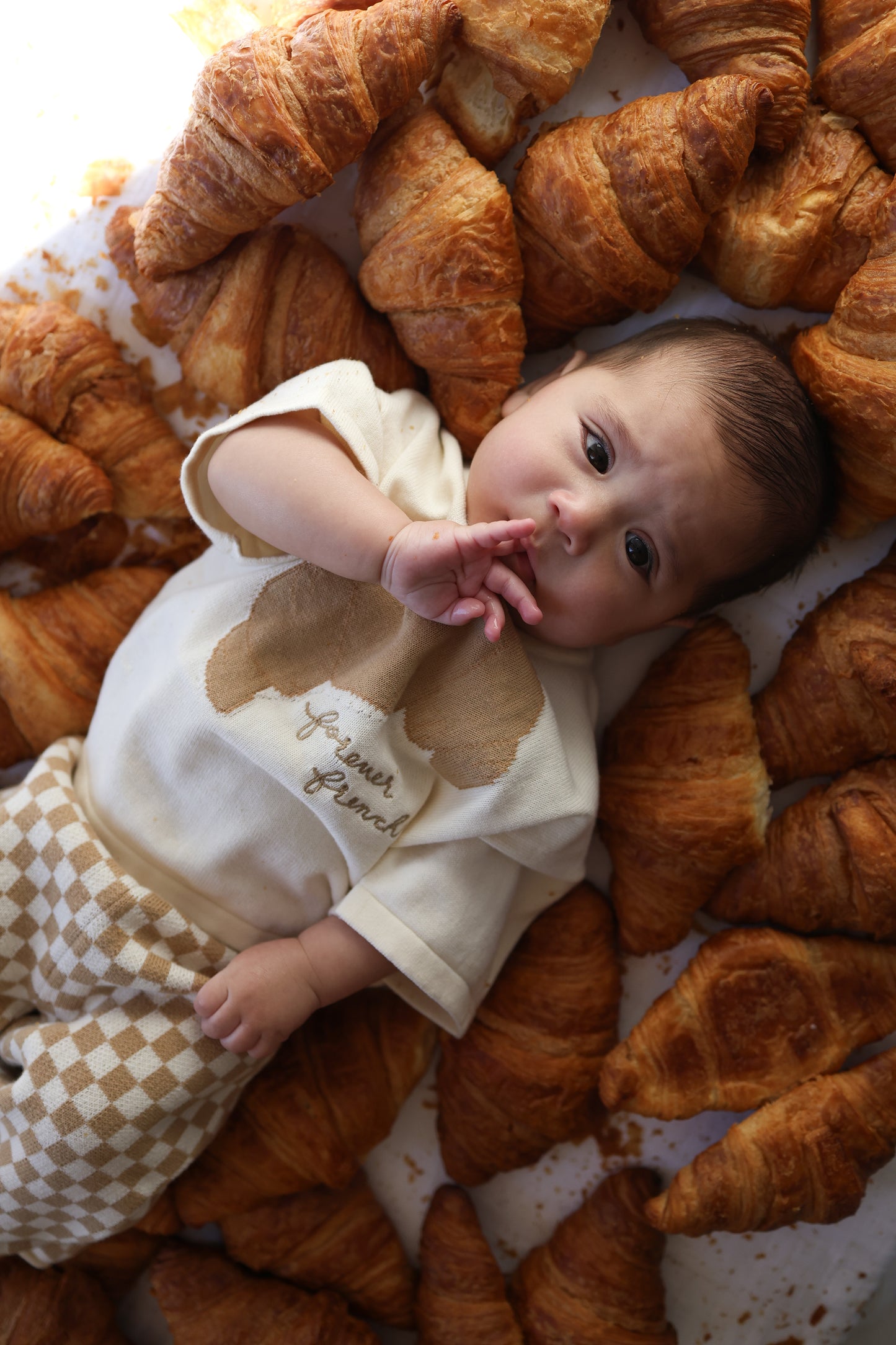 A baby wearing the forever french baby Knit Pant Set | Croissants lies surrounded by croissants, gazing upward with a hand near their mouth.
