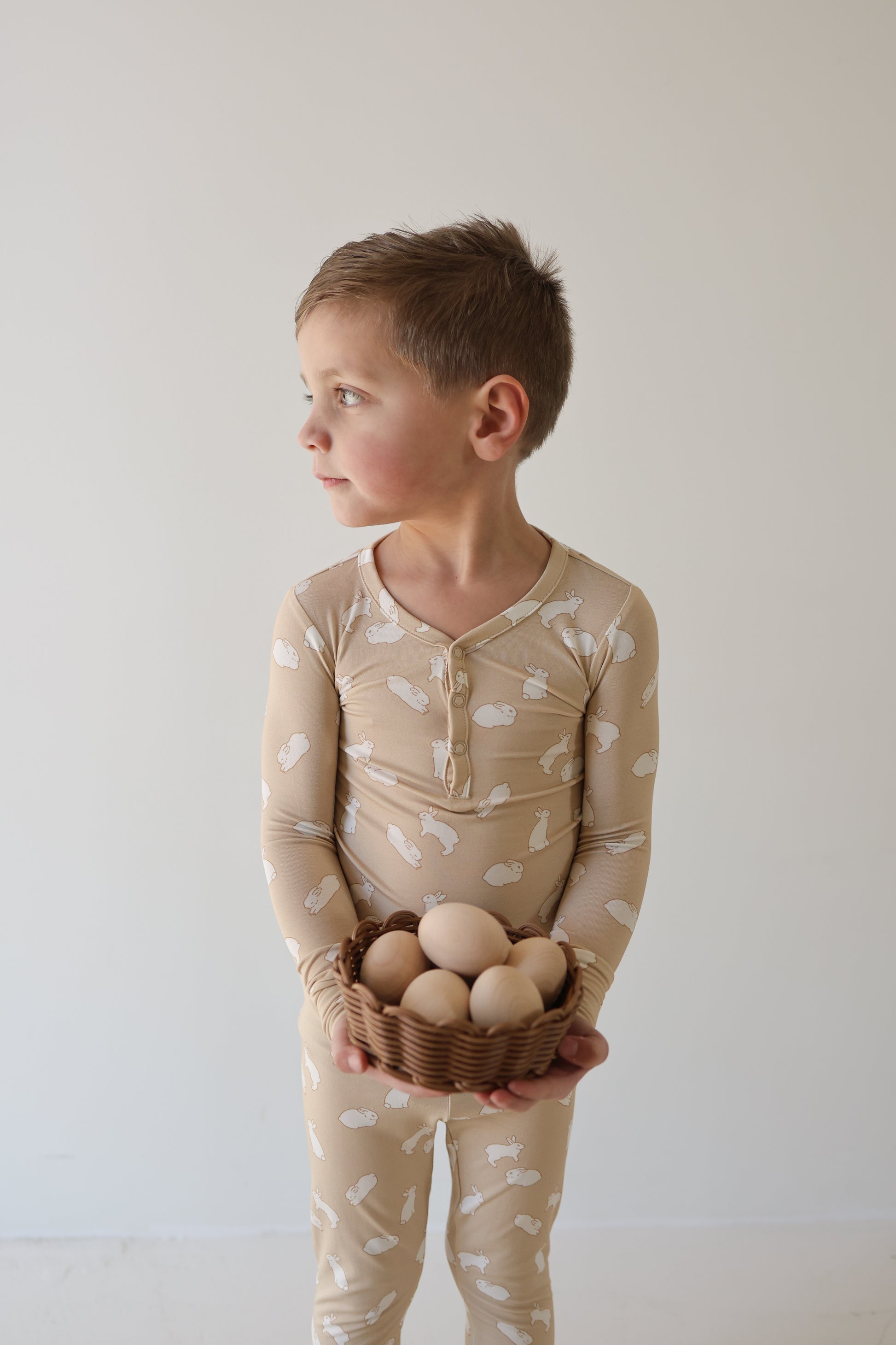 A young child in forever french baby Bamboo Two Piece Pajamas | Bunnies, featuring hypoallergenic fabric and white bunny prints, holds a small basket of brown eggs and looks to the side against a plain, light background.