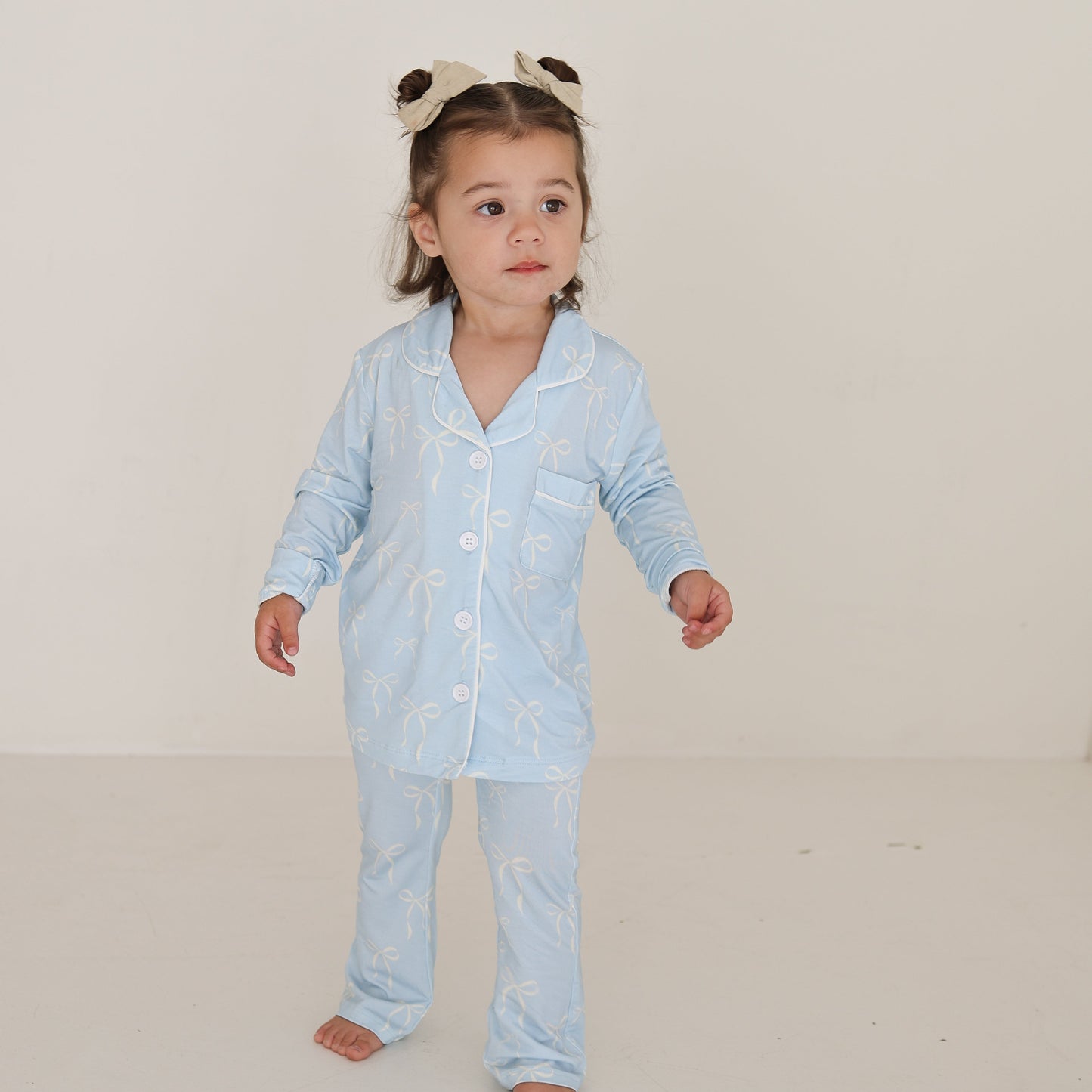 A young child in the forever french baby Girl’s Bamboo Button Up Lounge Set | Blue Bows stands against a white background, wearing hypo-allergenic, comfy bamboo clothing with a neutral expression and a cute bear-ear headband.