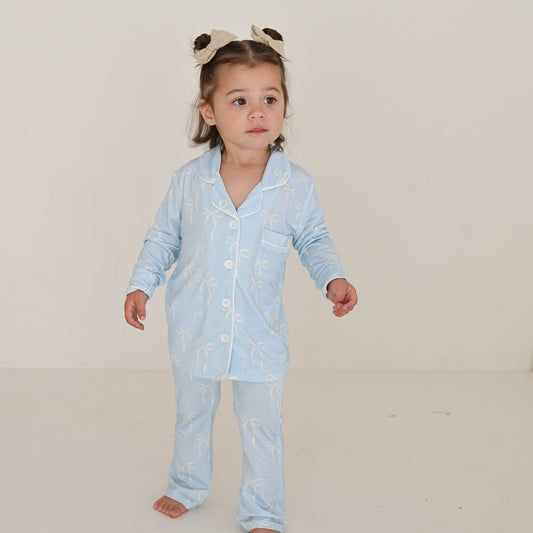 A young child in the forever french baby Girl’s Bamboo Button Up Lounge Set | Blue Bows stands against a white background, wearing hypo-allergenic, comfy bamboo clothing with a neutral expression and a cute bear-ear headband.