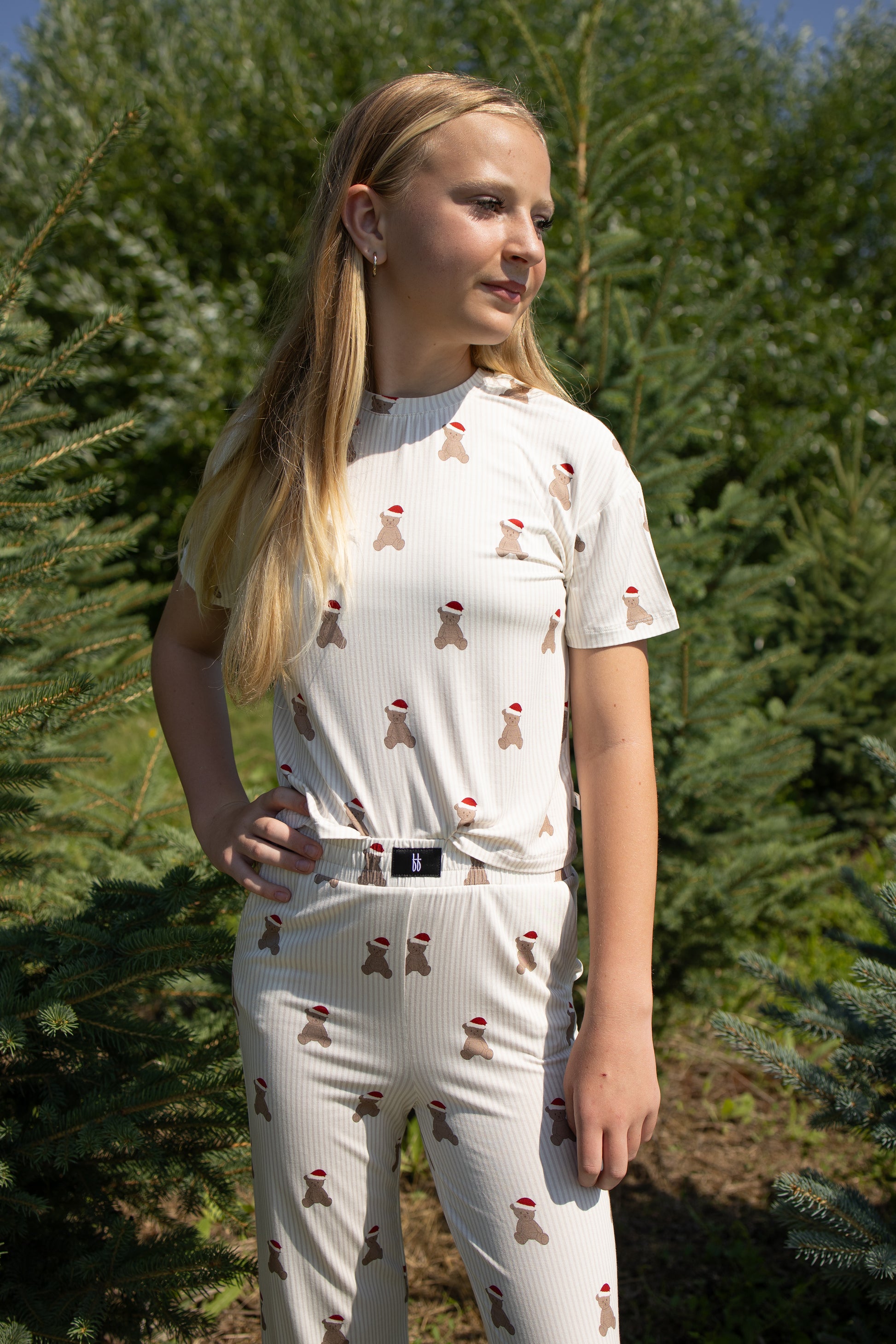 A girl with long blonde hair stands outdoors among pine trees, looking to the side with one hand on her hip. She is wearing the forever french baby Child Boxy Tee Lounge Set in the Santa Teddy print, a light-colored bamboo pajama set featuring small red and brown patterns.