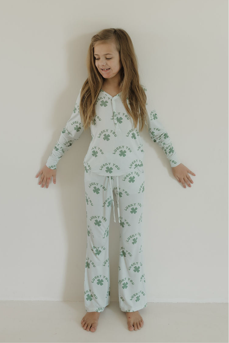 A young girl smiles barefoot against a white wall, wearing forever french baby’s Youth Flare Bamboo Pajamas | Lucky You—lightweight bamboo pajamas with green clovers and “Lucky You” text—for all-day breathable comfort.