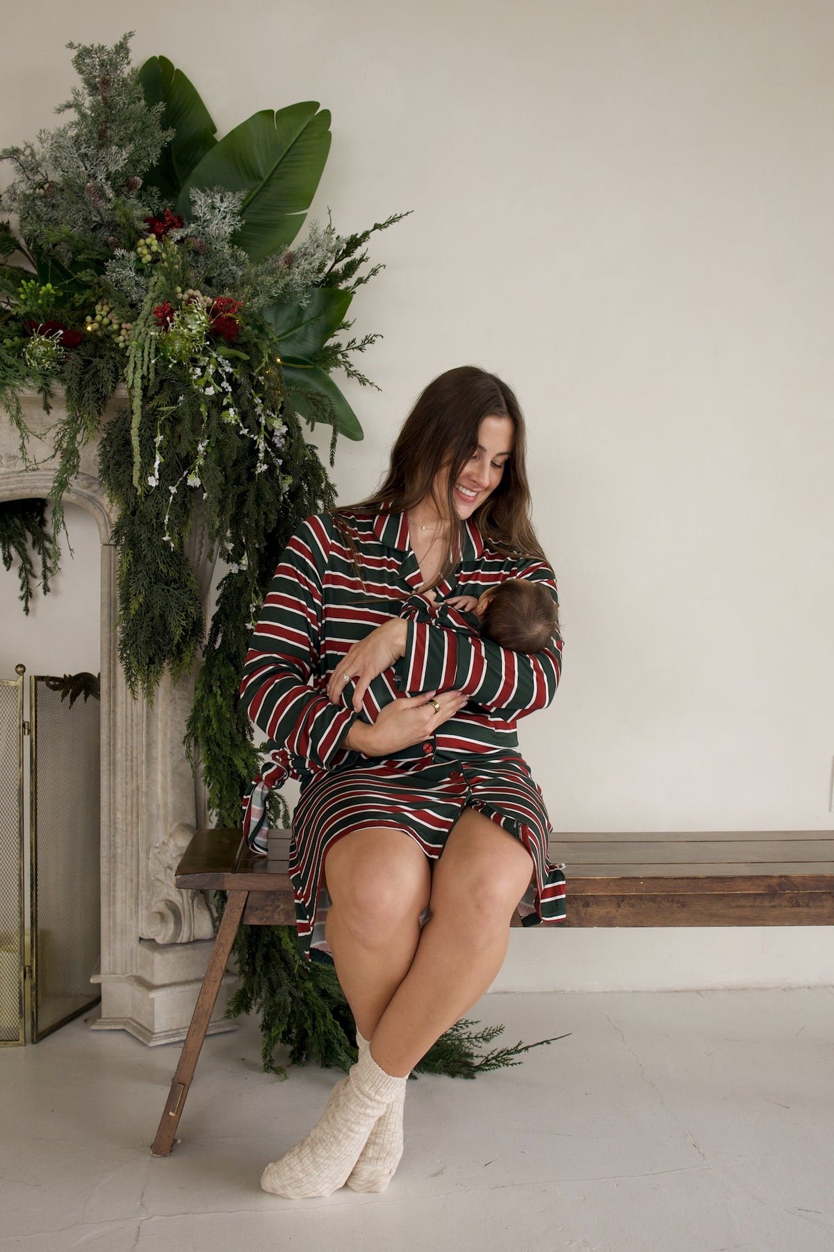 A woman wearing a red, green, and white Holiday Stripe Women’s Bamboo Sleeping Dress by forever french baby sits on a wooden bench while holding her baby.