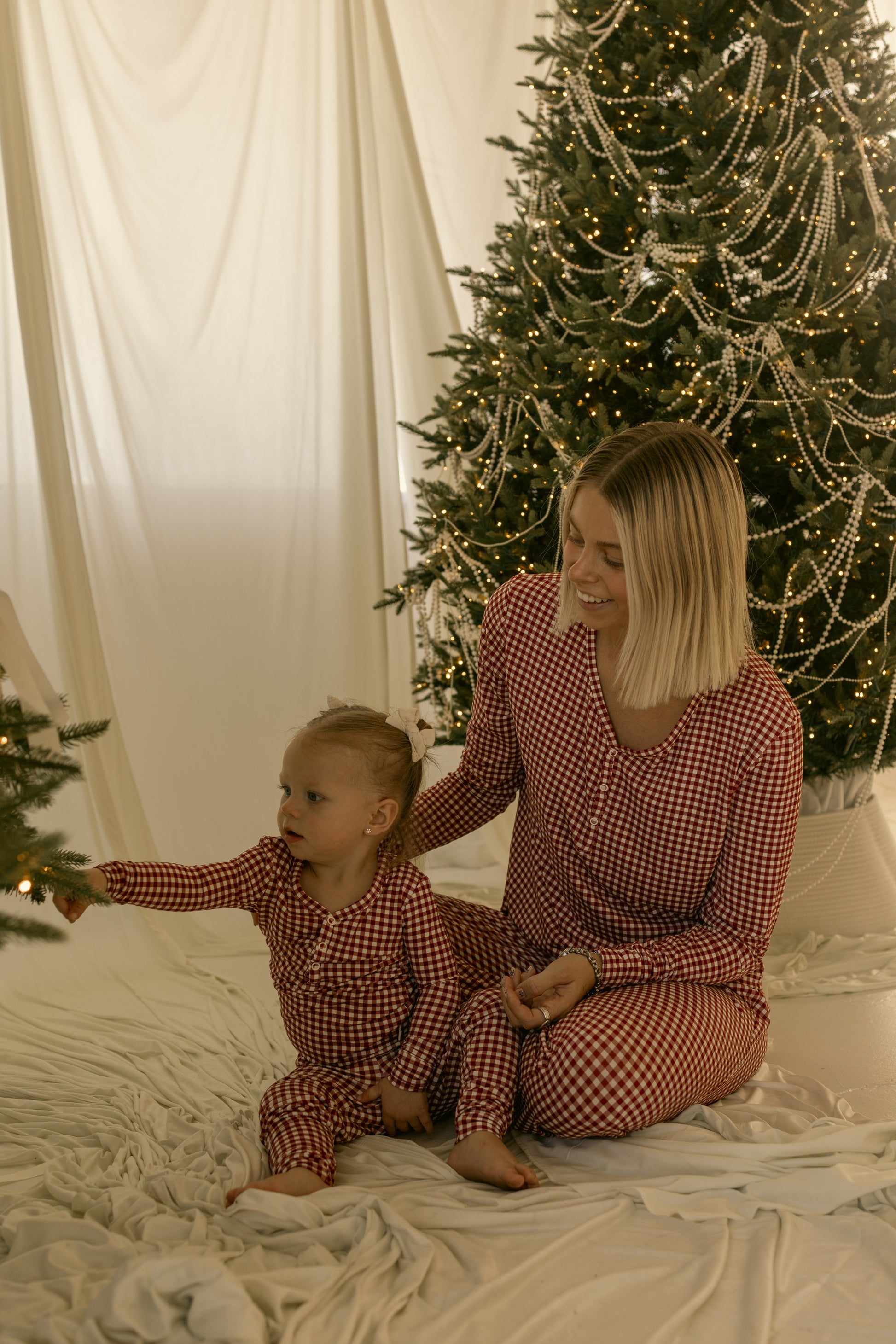 A woman and toddler in matching forever french baby Women's Bamboo Pajamas | Red Gingham sit on a white blanket by a Christmas tree with lights and pearl garlands; the toddler reaches for a branch.