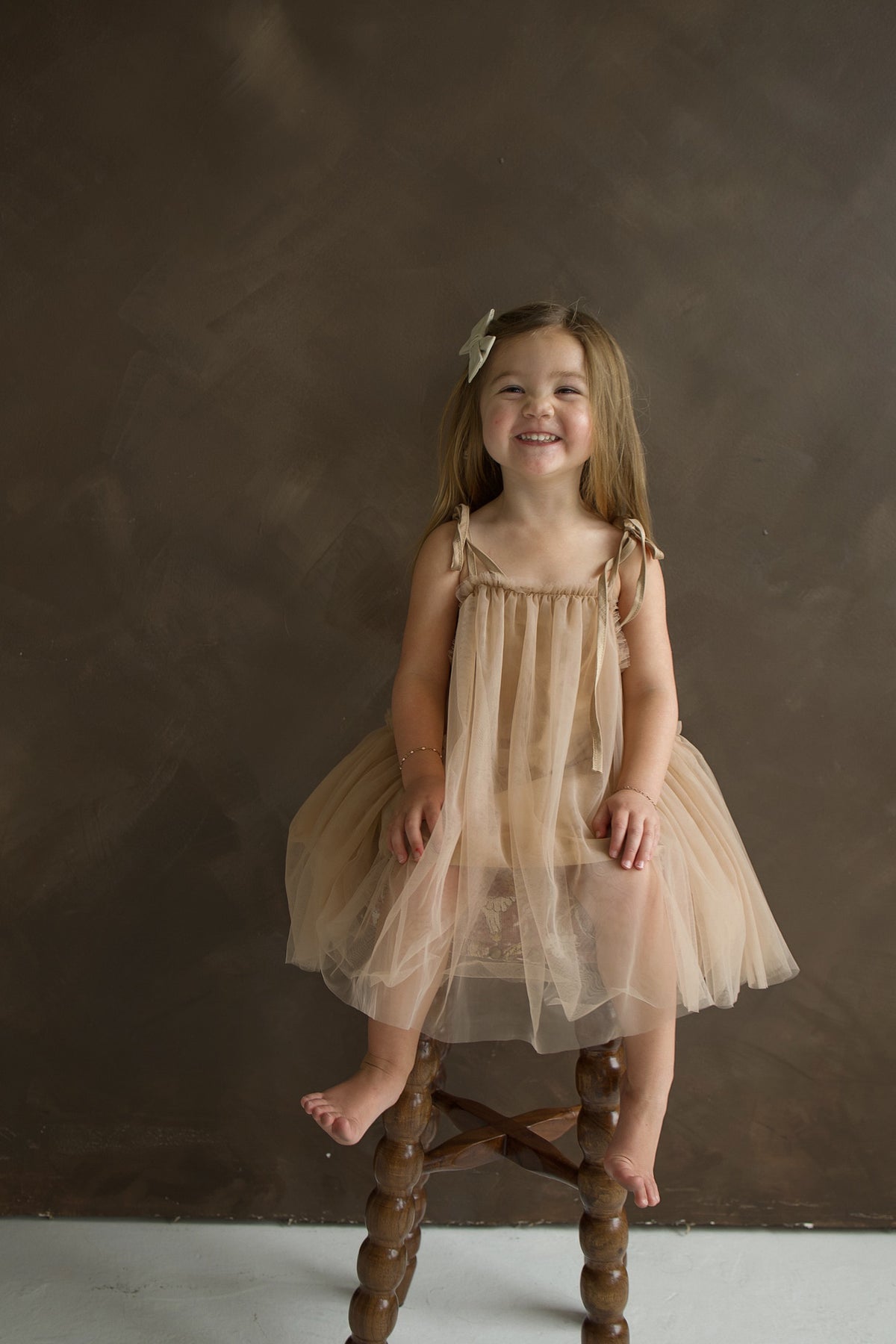 A smiling toddler wears a tan Tutu Dress | Teddy with bow straps while sitting on a wooden stool.
