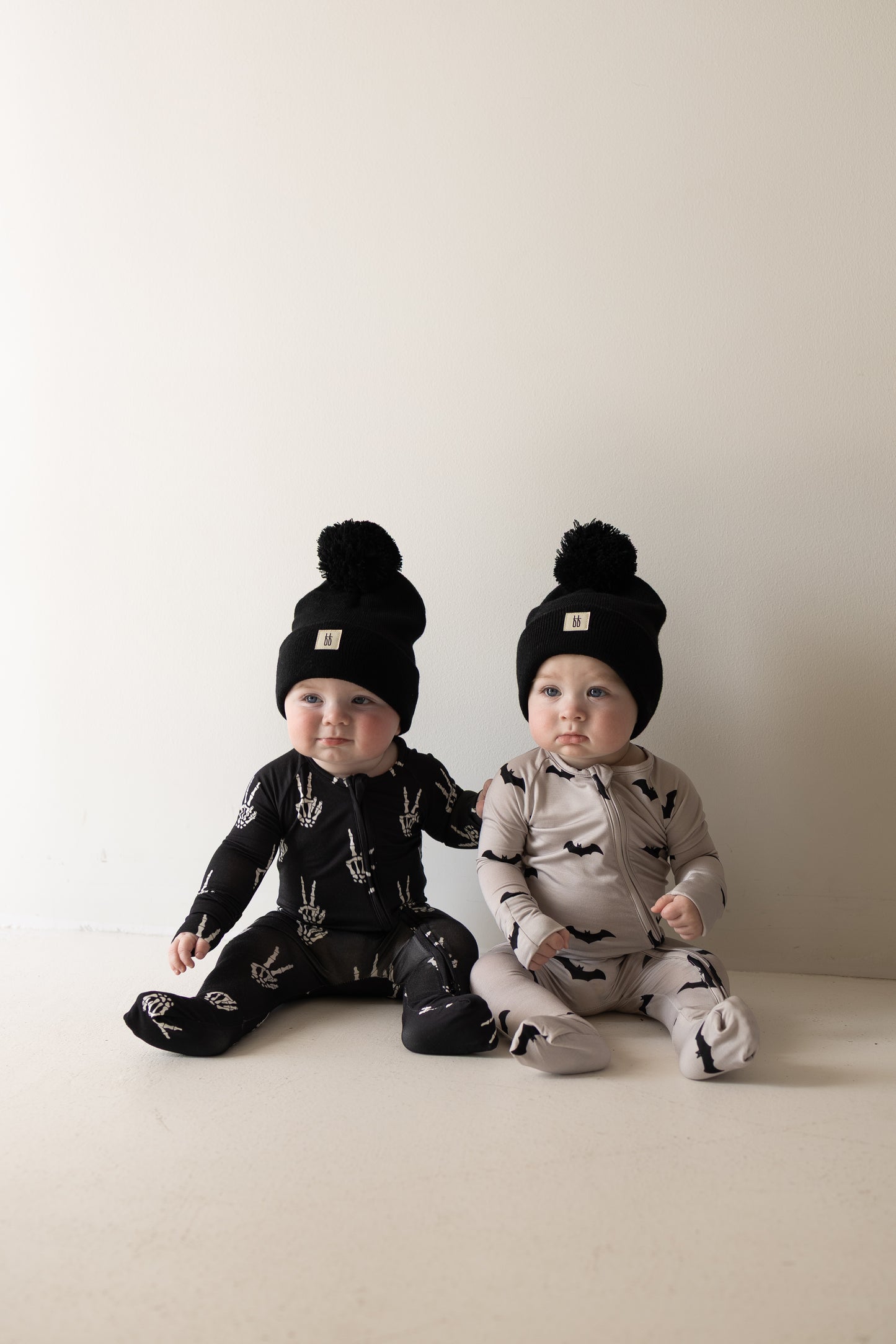 Two babies sit side by side wearing black pom-pom beanies—one dressed in forever french baby’s Bamboo Zip Pajamas | Midnight Skelly, featuring a black design with playful white skeleton hands, and the other in a light gray outfit adorned with black bats. Against a plain white background, their cozy pajamas crafted from hypo-allergenic bamboo viscose keep delicate skin comfortable and irritation-free.