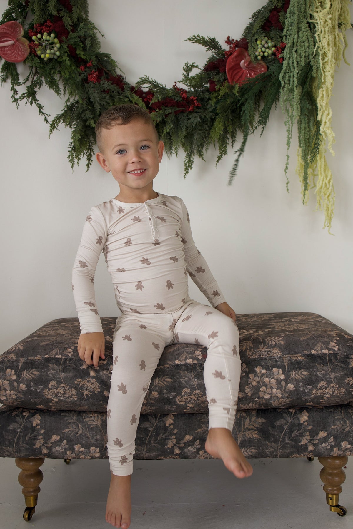 A smiling young boy wearing Bamboo Two Piece Pajamas | Gingersnap with a gingerbread man print, sitting on a patterned ottoman.