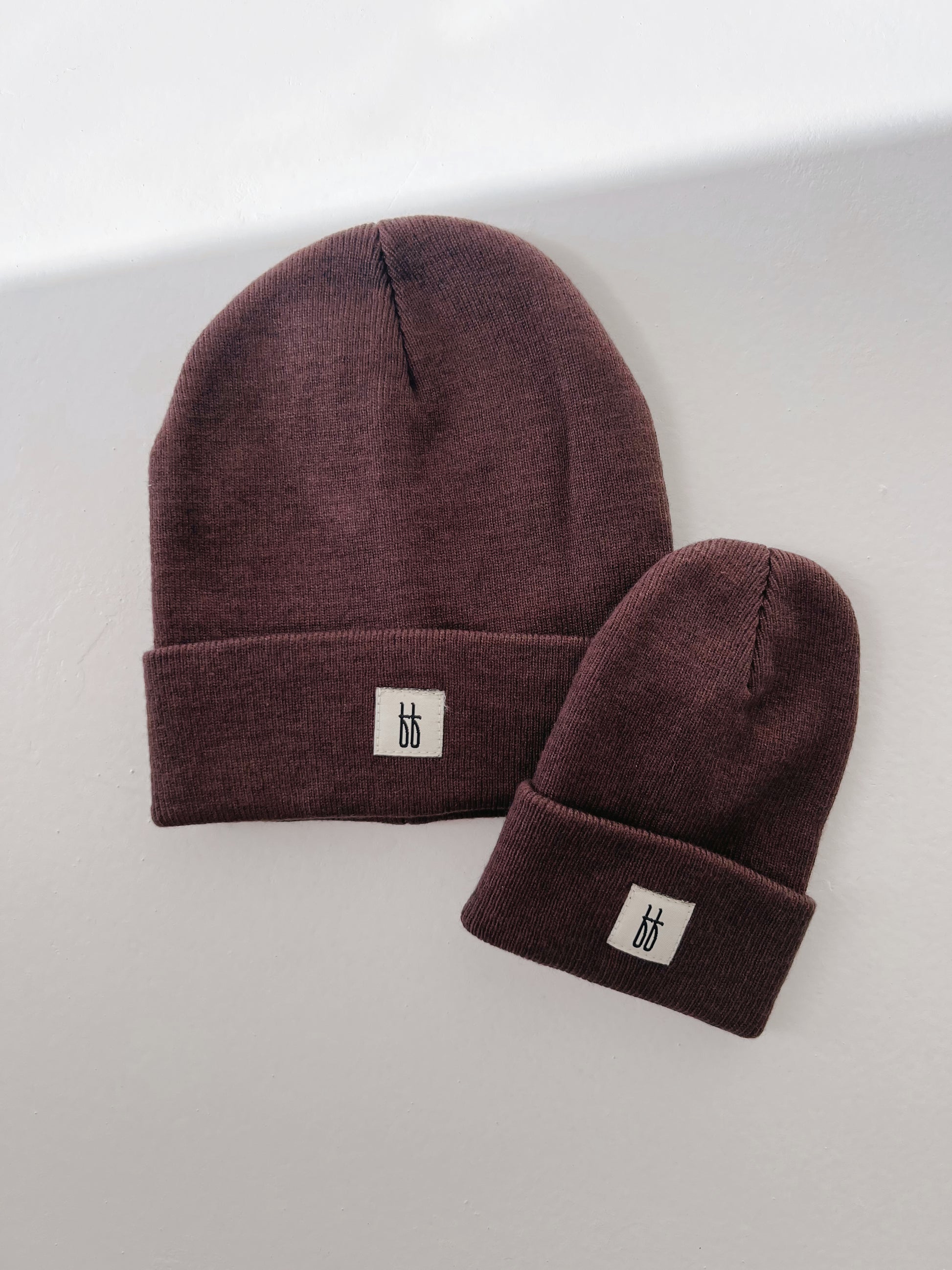 A photograph showing two coffee-colored ff knit beanies with a ribbed design on a white surface. The larger ff Knit Beanie by forever french baby is for an adult, while the smaller one, perfect for a child, is also from forever french baby. Both feature a small white sewn-on label with the same design logo on the front.
