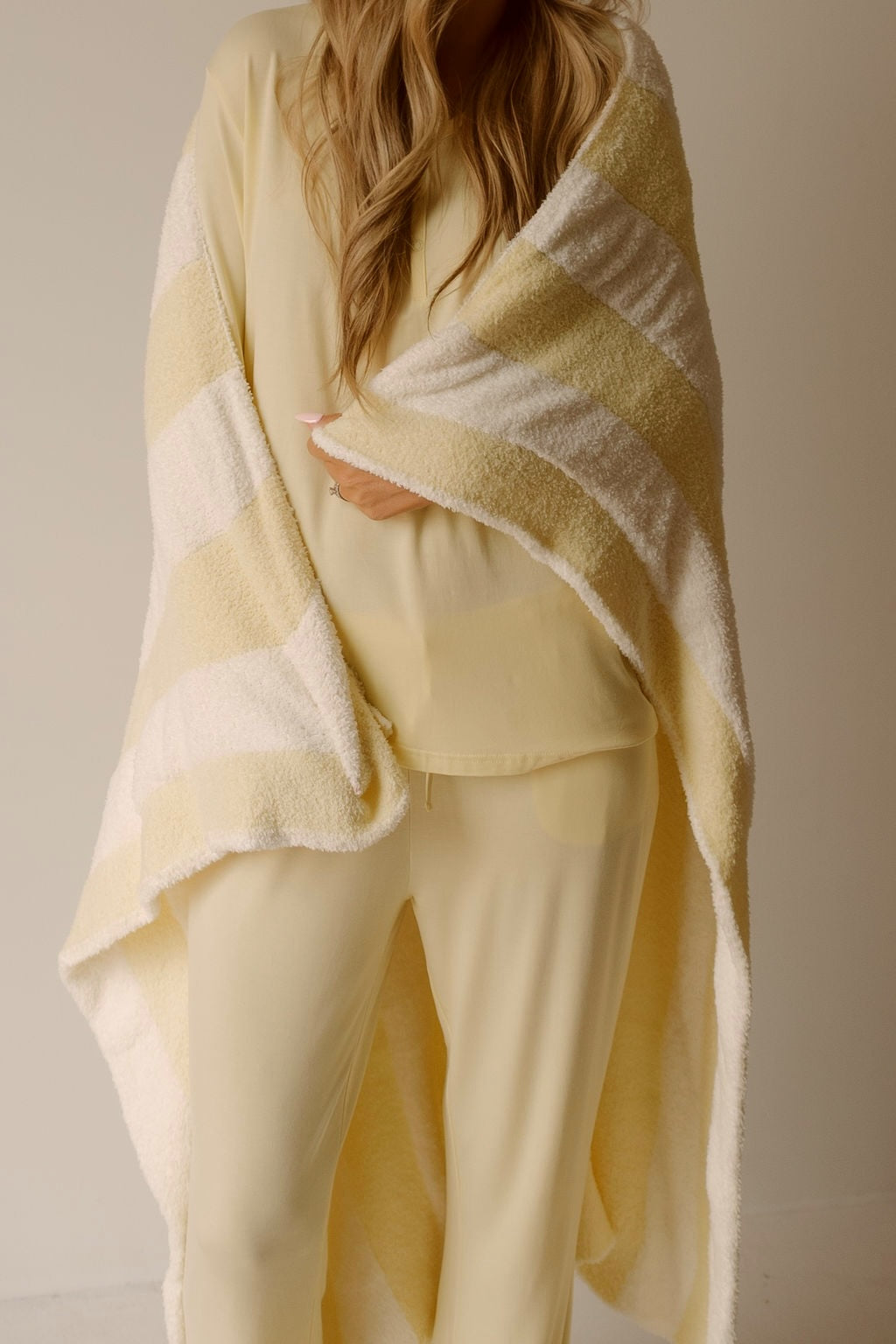 A person with long, wavy hair, wearing light yellow pajamas, wraps themselves in the forever french baby Plush Blanket | Sunny Stripe—a soft, OEKO-TEX certified blanket featuring yellow and white stripes. Their face is not visible.
