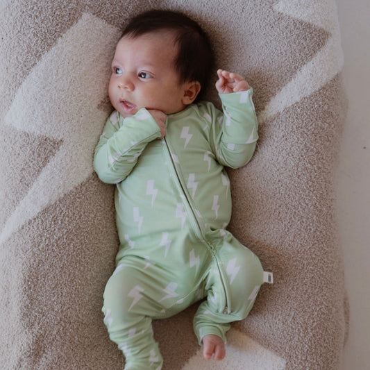 A baby in forever french baby's Bamboo Zip Pajamas | Flash—a mint green onesie with white lightning bolts—lies on a soft, beige cushion with white geometric patterns, gazing to the side.