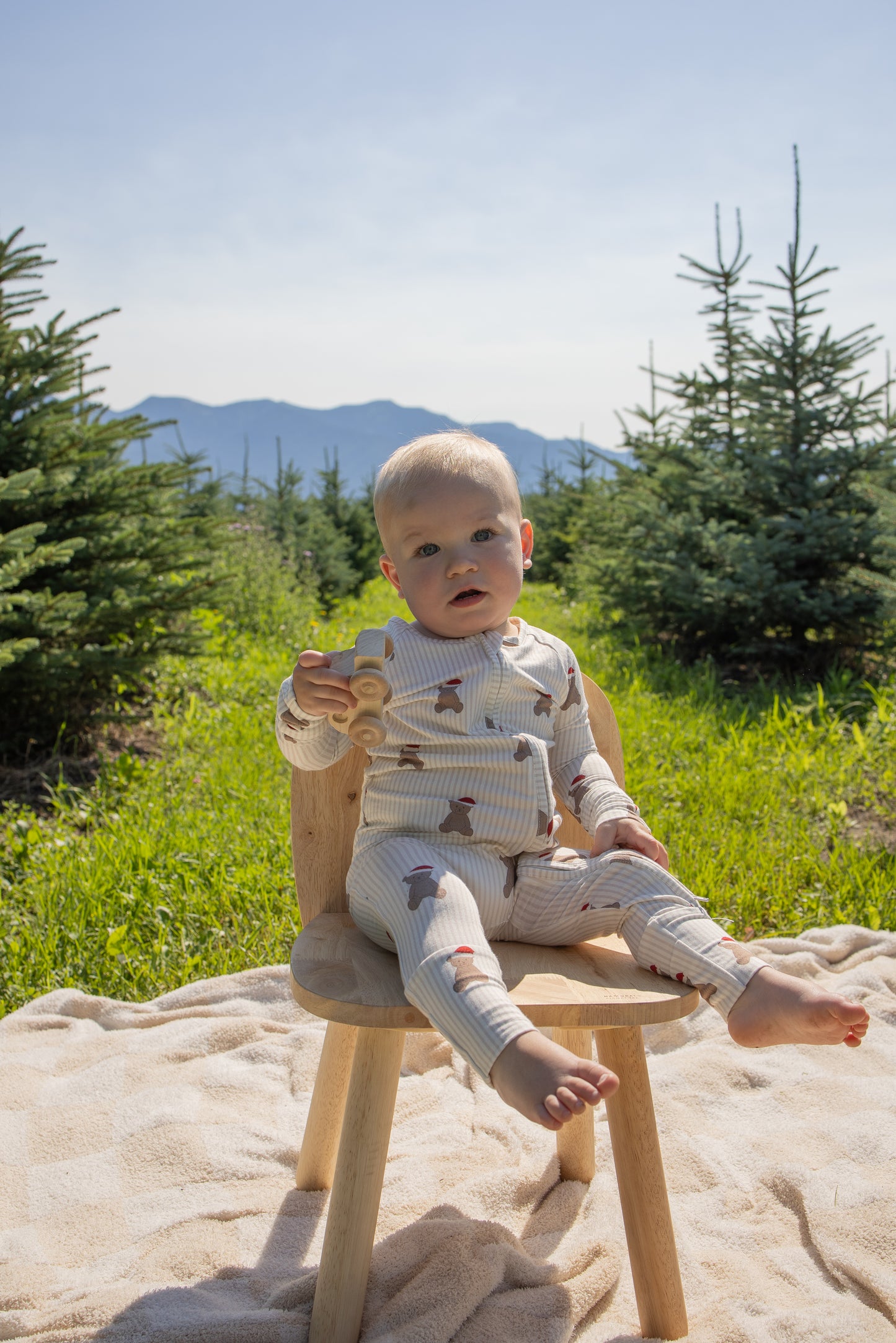 A baby dressed in the forever french baby Bamboo Zip Pajamas | Santa Teddy sits on a wooden chair outdoors, holding a wooden toy truck, surrounded by small pine trees and mountains in the background with sunlight streaming in.