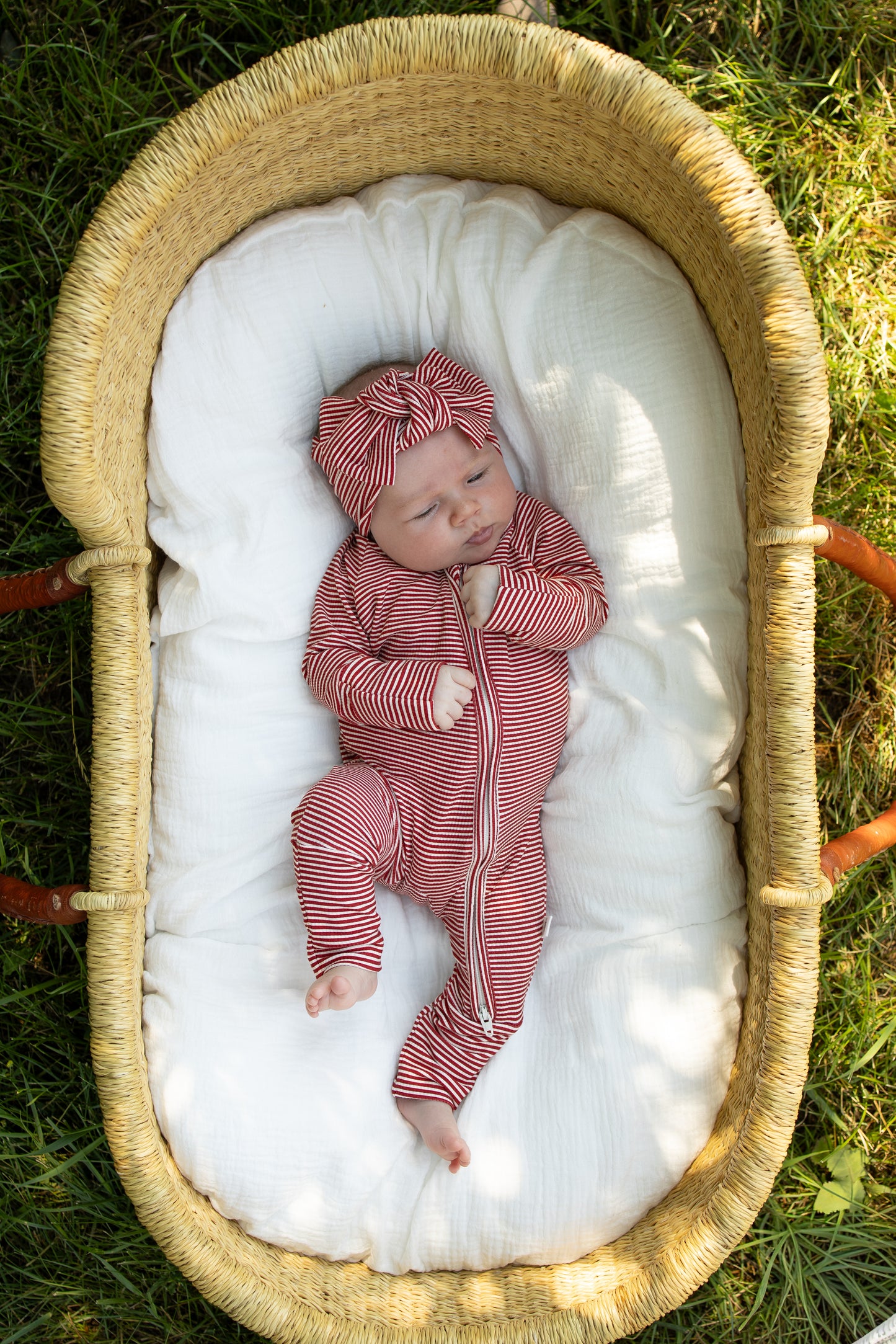 A baby wearing the forever french baby Bamboo Zip Pajamas in Peppermint Stripe Ribbed with a matching headband lies in a wicker basket on a white blanket outdoors, resting on the grass and looking up with one hand near their face.
