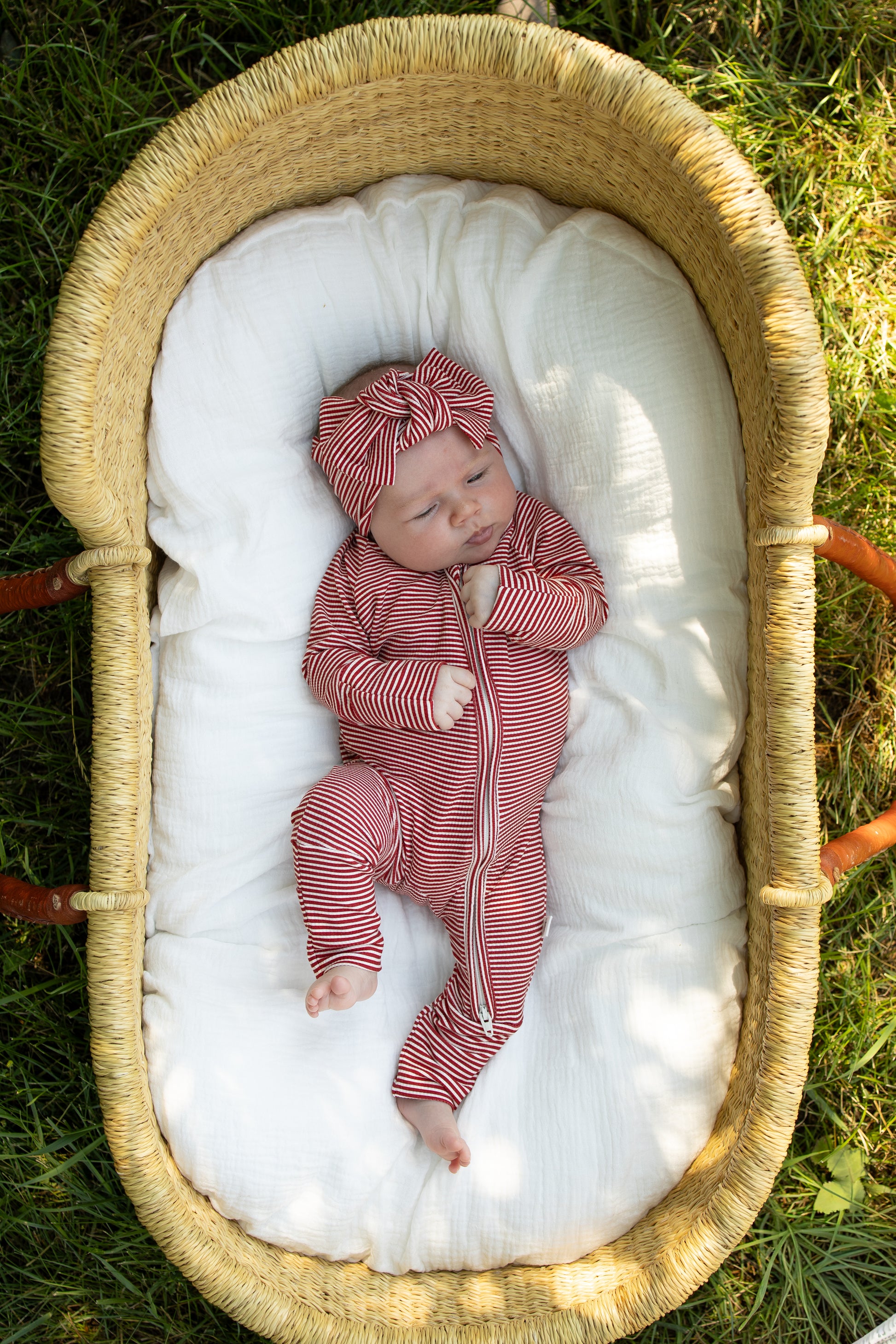 A baby wearing the forever french baby Bamboo Zip Pajamas in Peppermint Stripe Ribbed with a matching headband lies in a wicker basket on a white blanket outdoors, resting on the grass and looking up with one hand near their face.