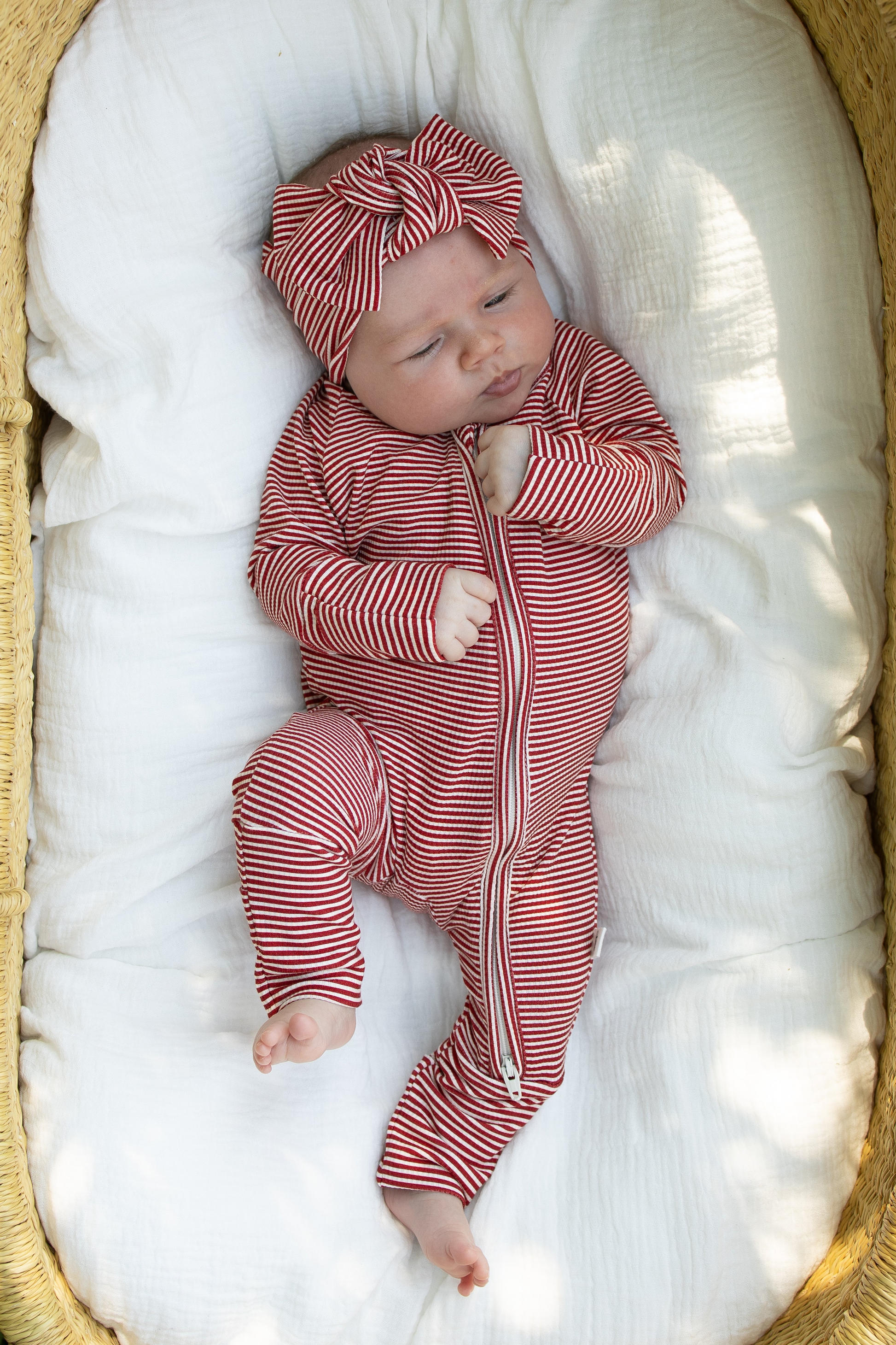A baby wearing forever french baby's Bamboo Zip Pajamas in Peppermint Stripe Ribbed, along with a matching headband, lies on a white blanket in a woven basket, gazing upward with one hand near their face.