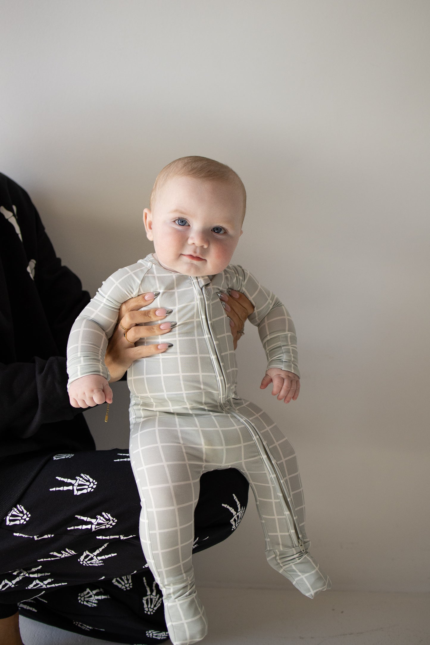 Clad in the forever french baby Bamboo Zip Pajamas in the Foggy Grid pattern, a baby—dressed in soft, breathable sleepwear—is held up by an adult who is mostly out of frame. The baby gazes at the camera with a neutral expression.
