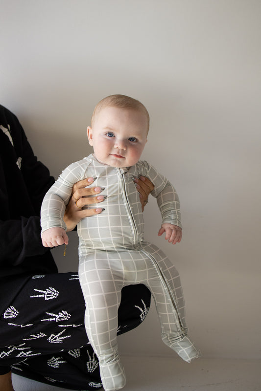 Clad in the forever french baby Bamboo Zip Pajamas in the Foggy Grid pattern, a baby—dressed in soft, breathable sleepwear—is held up by an adult who is mostly out of frame. The baby gazes at the camera with a neutral expression.
