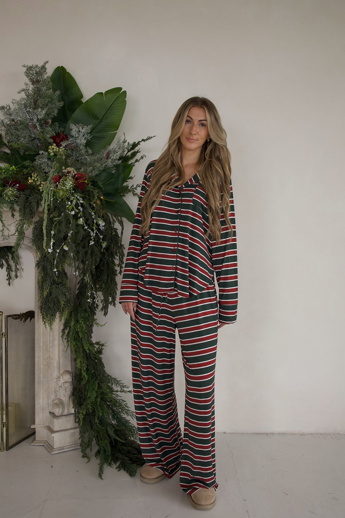 A woman wears a Women’s Bamboo Button Up Lounge Set | Holiday Stripe featuring a long-sleeved button-up shirt and wide-leg lounge pants in a red, green, and white horizontal stripe pattern.