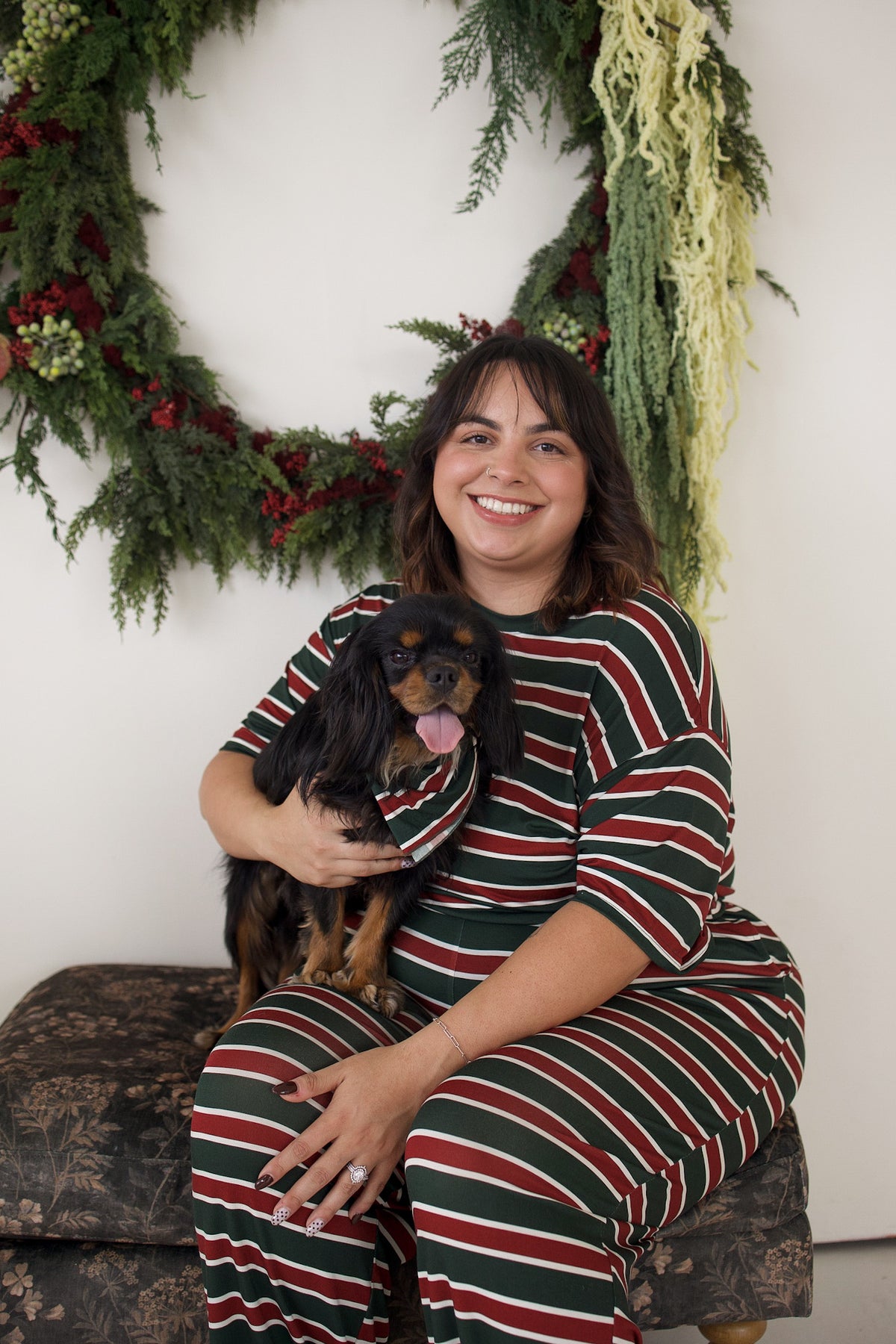 A smiling woman wearing a red, green, and white Women’s Boxy Tee Lounge Set | Holiday Stripe holds a small dog in a striped bandana in front of a holiday wreath.