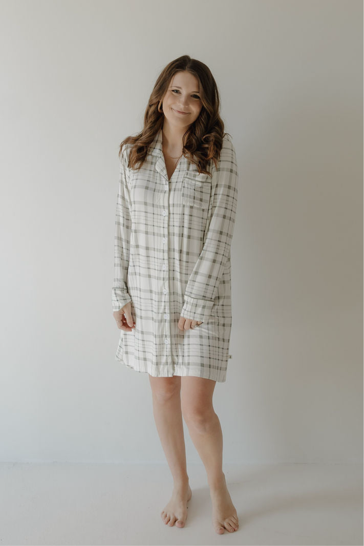 A woman with long brown hair smiles barefoot in a light-filled room, wearing the forever french baby Women's Bamboo Sleeping Dress | Picnic Plaid—a breathable white and gray plaid long-sleeve shirt dress—standing by a plain, light wall.