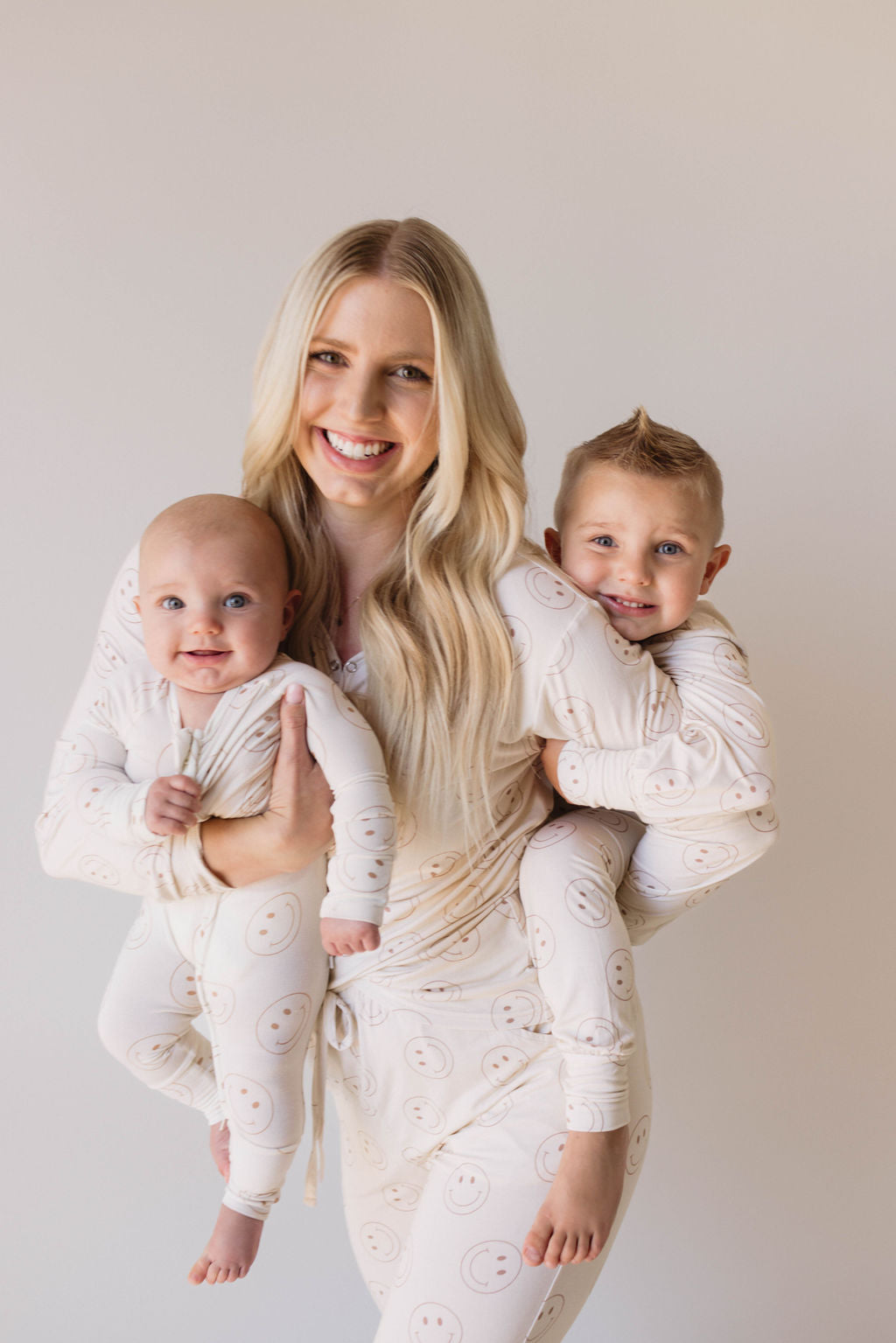 A smiling woman with long blonde hair is holding two young children, each on one arm. All three are donning matching white outfits adorned with smiley face patterns, crafted from soft bamboo viscose. The backdrop of a plain white wall perfectly showcases their cozy and stylish Women's Bamboo Pajamas from the "Just Smile" collection by forever french baby.