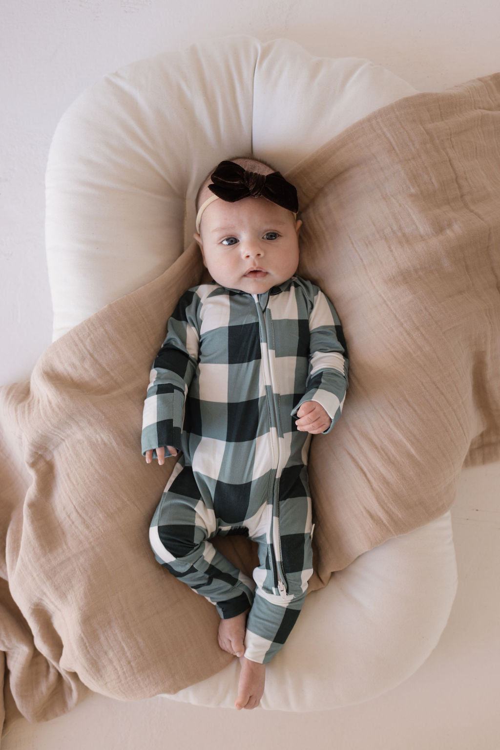 A baby lies on a cushioned surface, swaddled in a light brown blanket and dressed in the Evergreen Gingham Bamboo Zip Pajamas by forever french baby. A black headband with a bow adds a touch of comfort and style to the outfit.