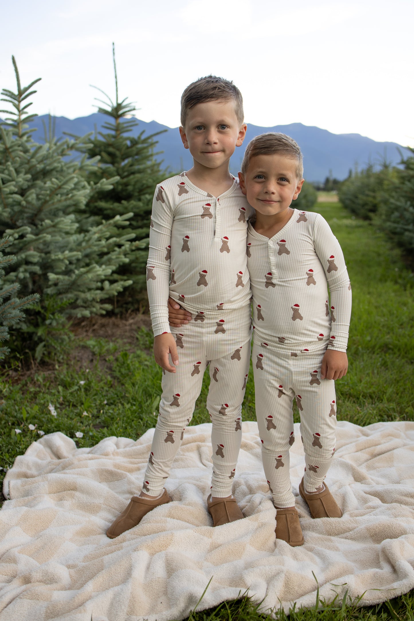 Two young boys in matching forever french baby Bamboo Two Piece Pajamas | Santa Teddy stand together on a blanket outdoors among pine trees, with mountains and sky in the background. Their cozy hypo-allergenic pajamas keep them comfortable as they hug and smile at the camera.