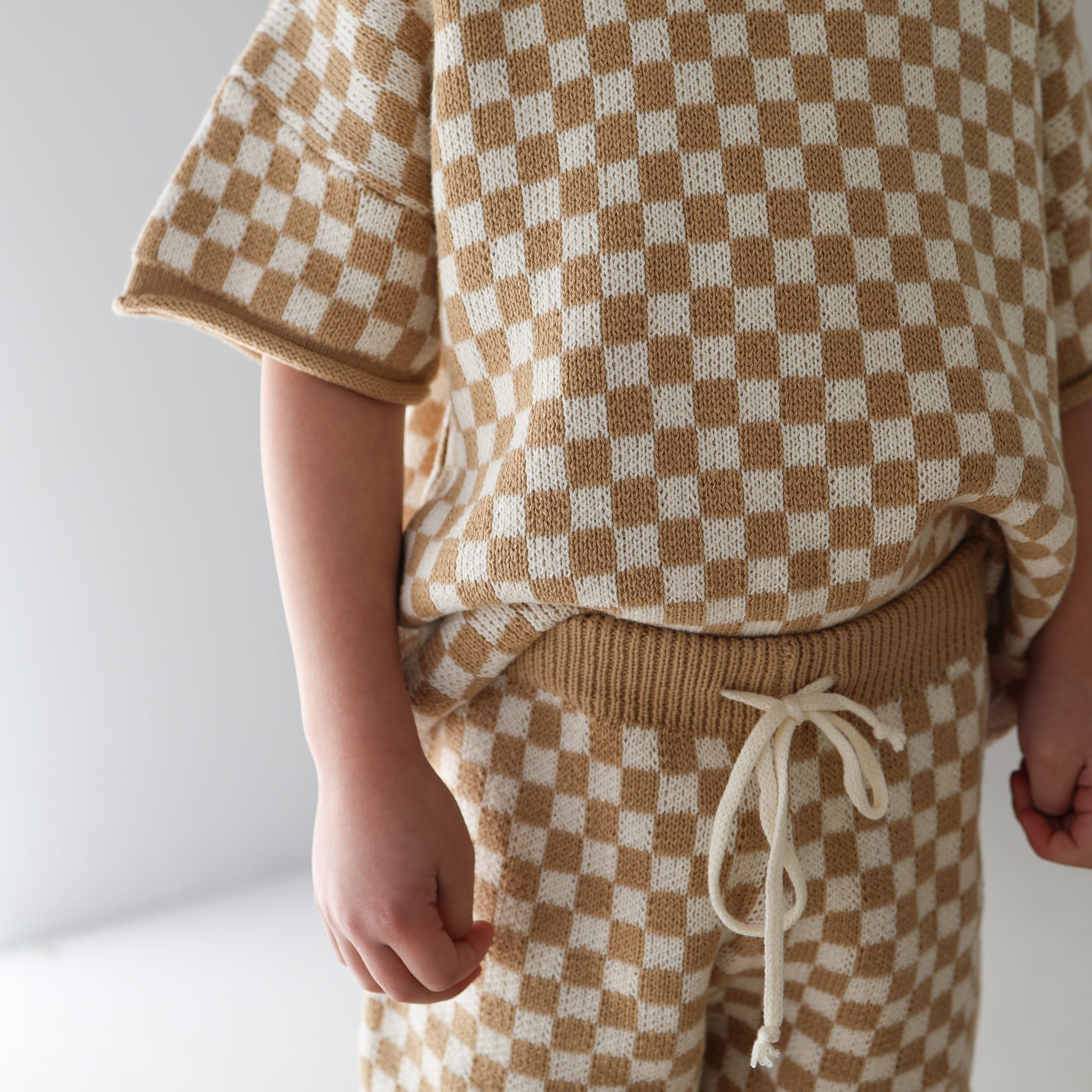 A child wears forever french baby's Knit Short Set | Latte Check—beige and white knit top with matching drawstring shorts—standing indoors against a plain light background.