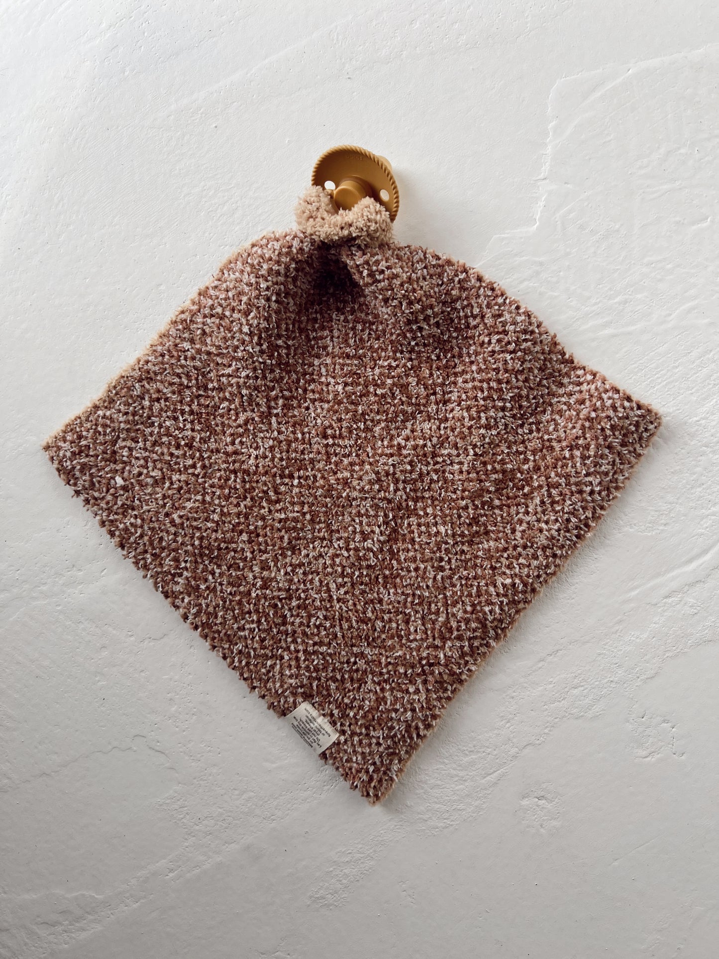 A charming brown textured knit cloth from the Gingerbread collection, named "Lovey" by forever french baby, hangs from a gold hook on a white wall. This square-shaped piece, made of microfiber feather yarn, includes a small white tag in one corner.